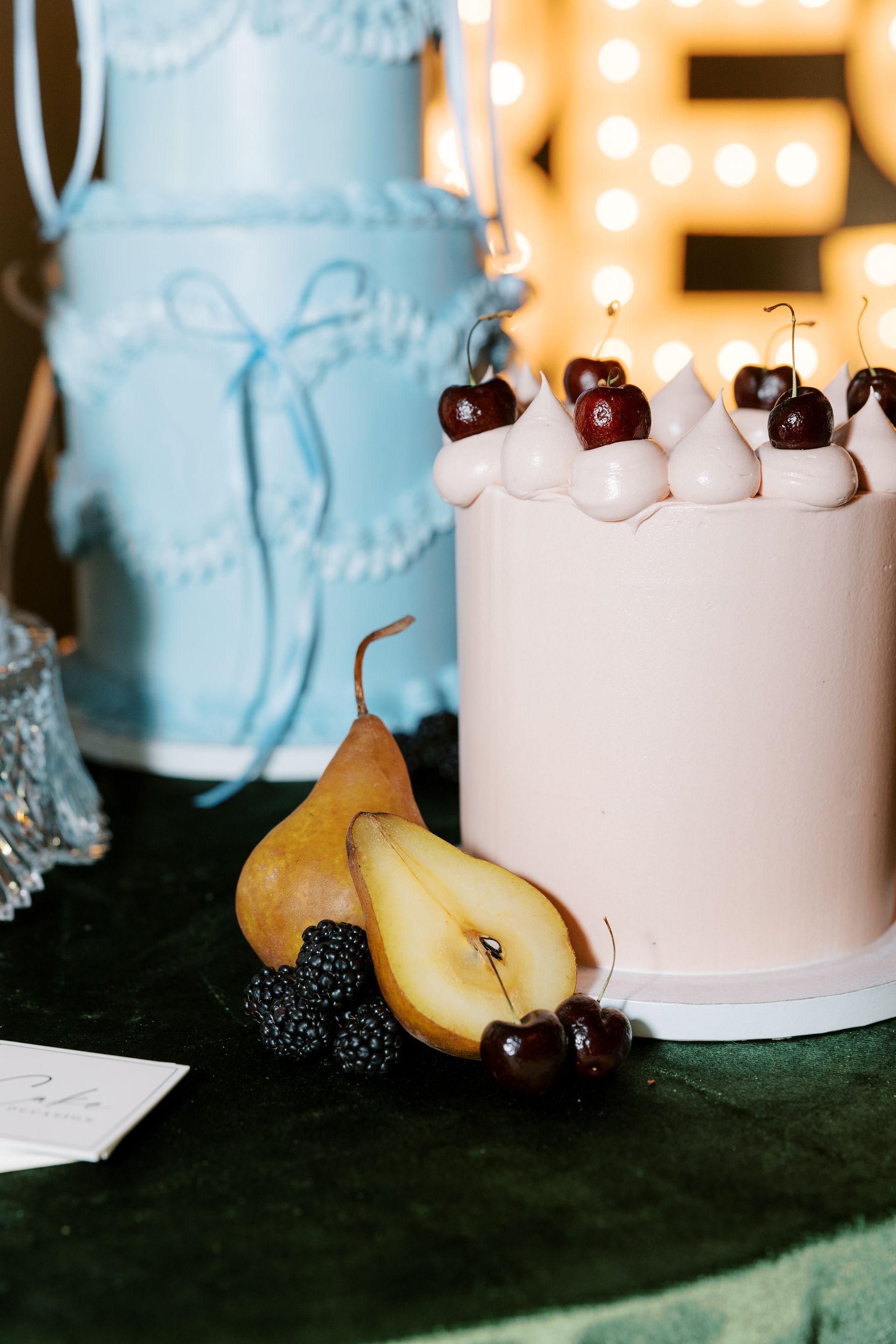 Pink cake with cherries and pears on a green surface; blue cake in background.