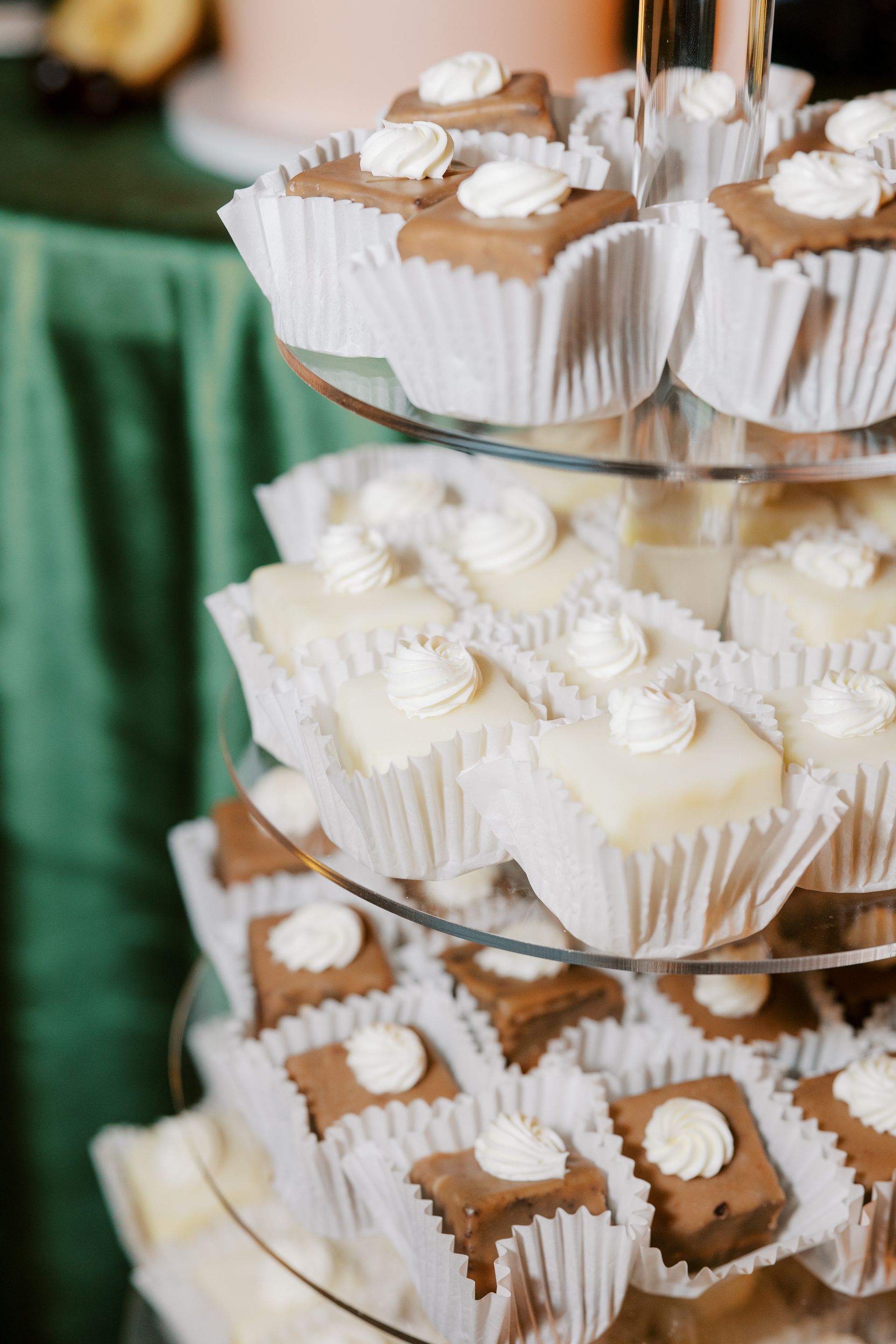 Tiered dessert display with square cakes, white and brown, topped with white frosting, on a table with a green tablecloth.