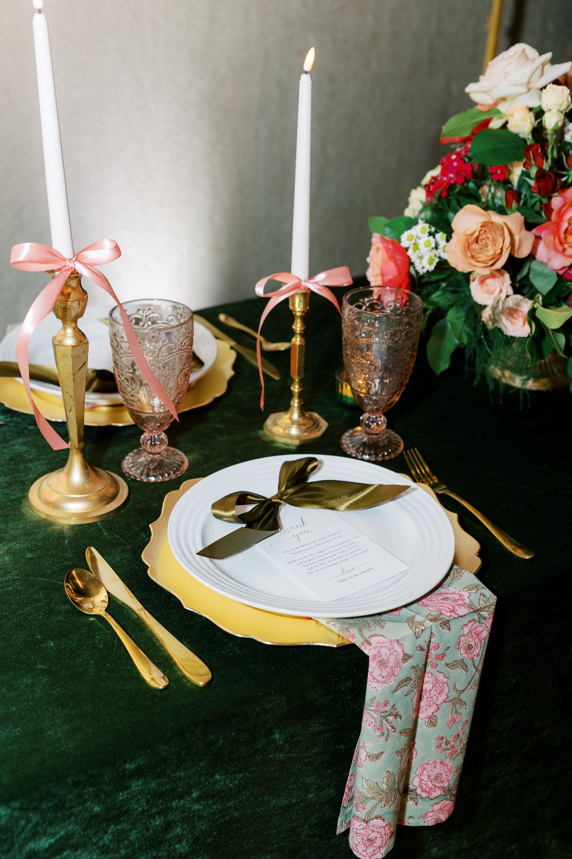 Elegant table setting with gold accents, pink ribbon, floral centerpiece, and green tablecloth.