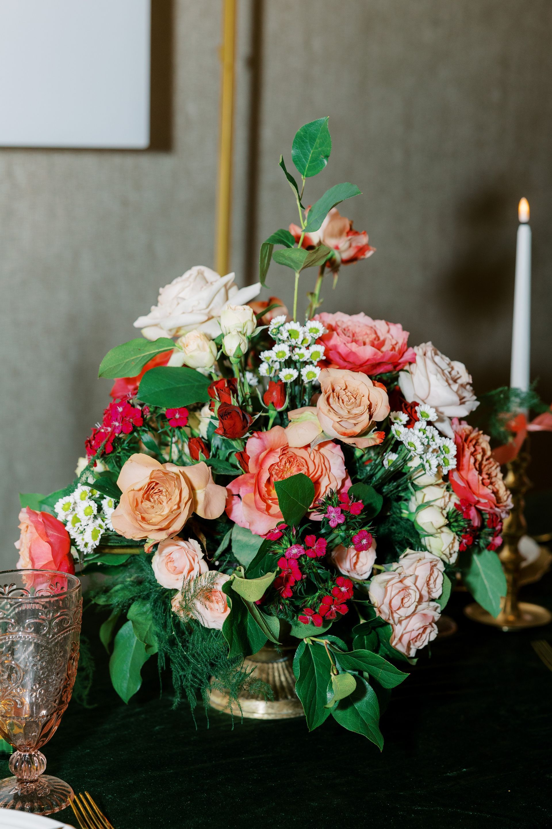 Floral centerpiece with roses, greens, and small white flowers on a table with candle.