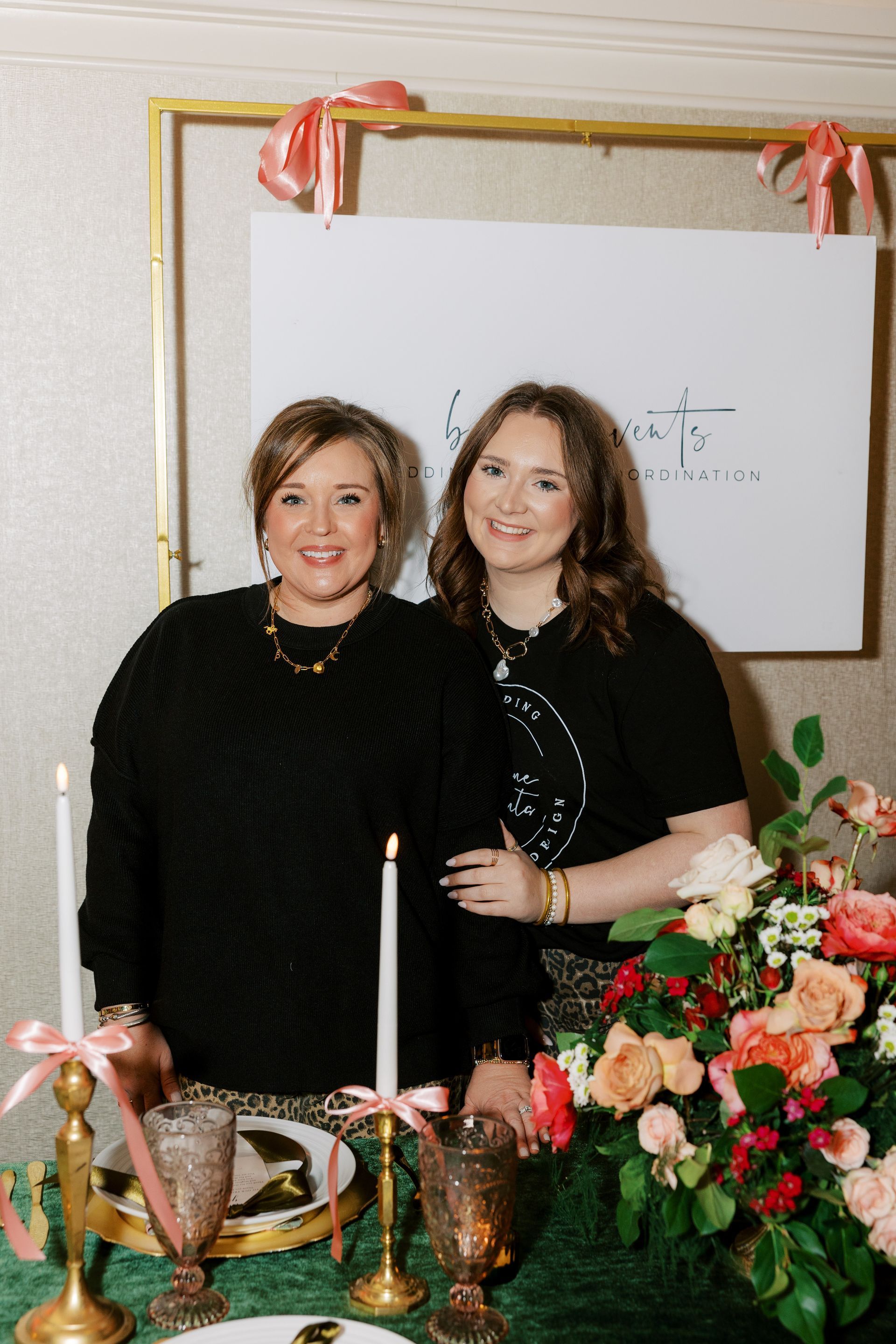 Two women pose near a decorated table with flowers and a sign.