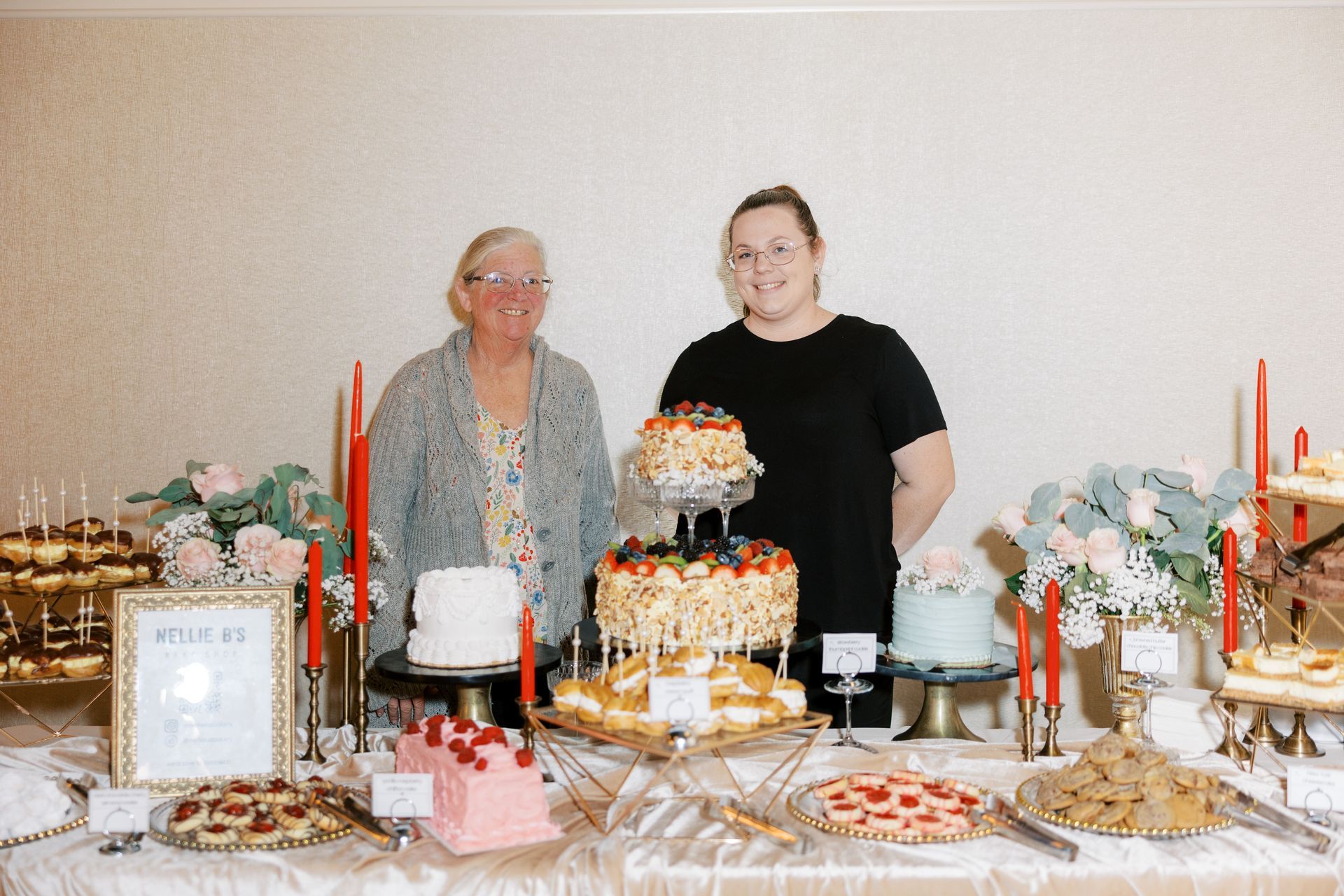 Two women stand behind a dessert table with various cakes, tarts, and pastries, indoors with flowers and candles.
