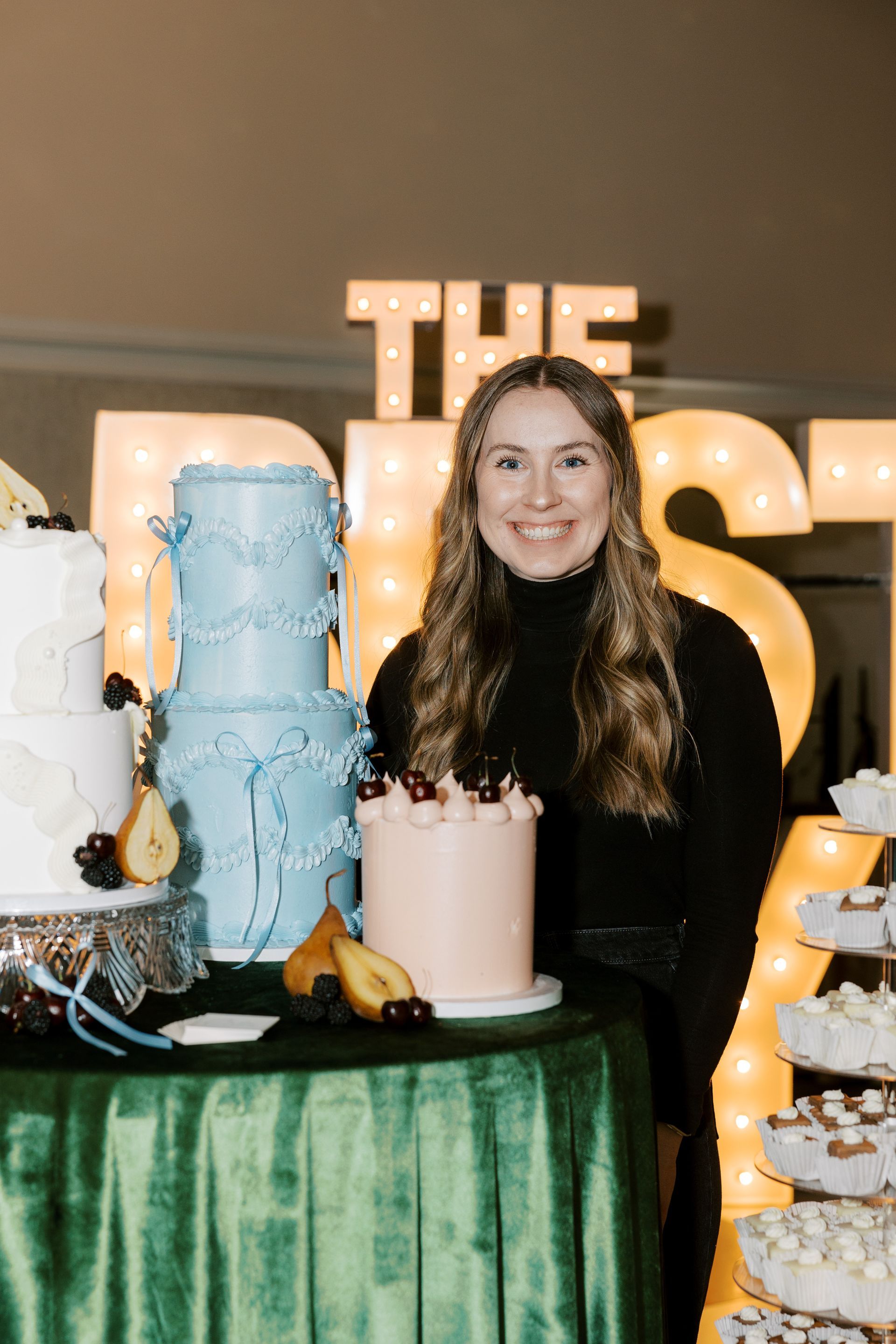 Woman smiles near three decorated cakes, green table, and 