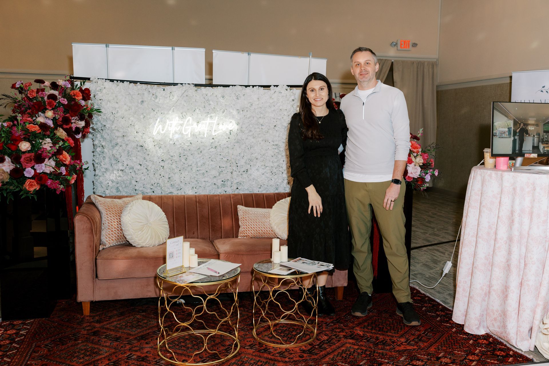 Couple poses by a pink couch and floral backdrop at an event; tables with products are in the foreground.
