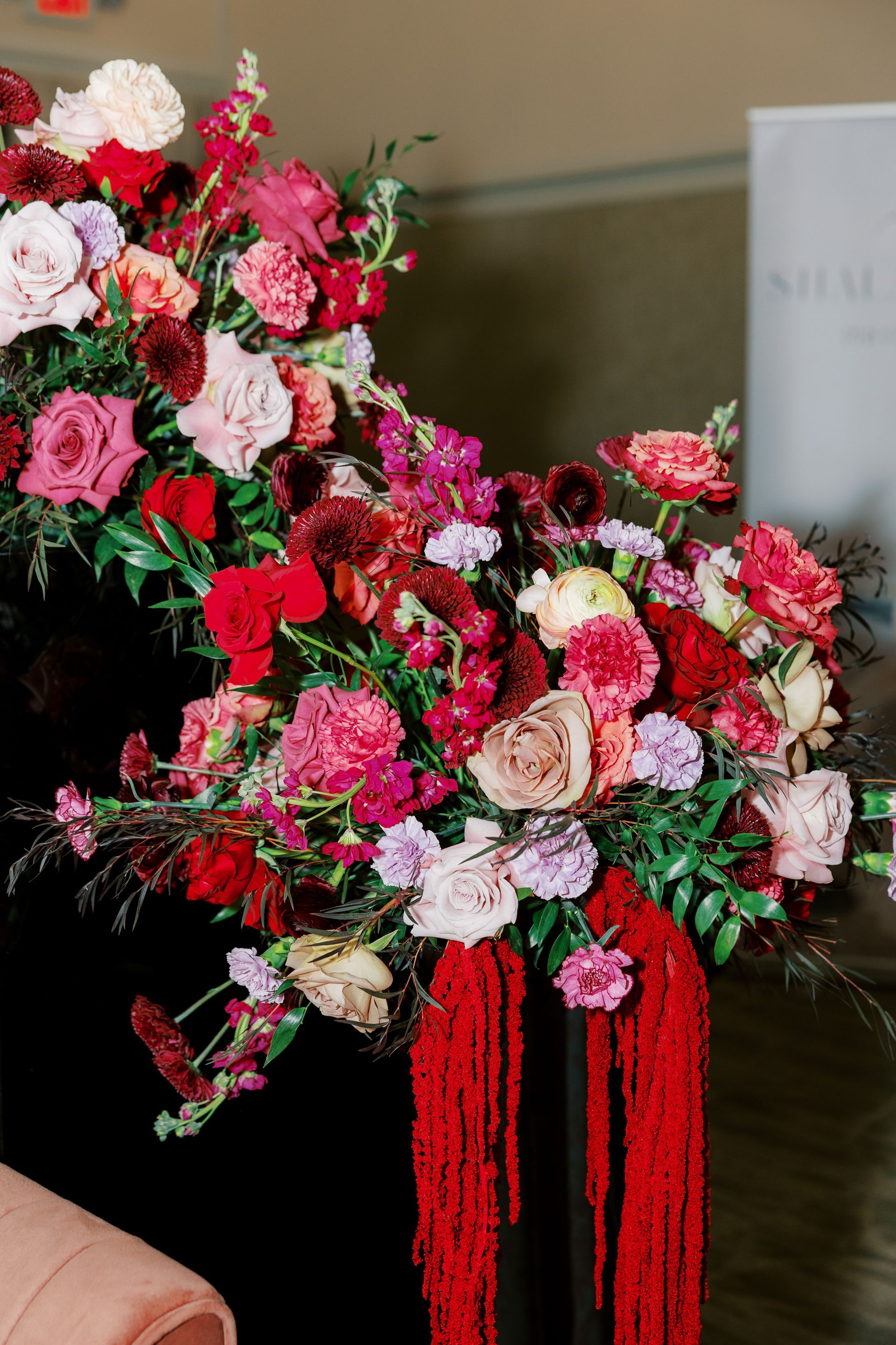 Arrangement of red, pink, and cream flowers spilling from a dark surface with red fringe, indoors.