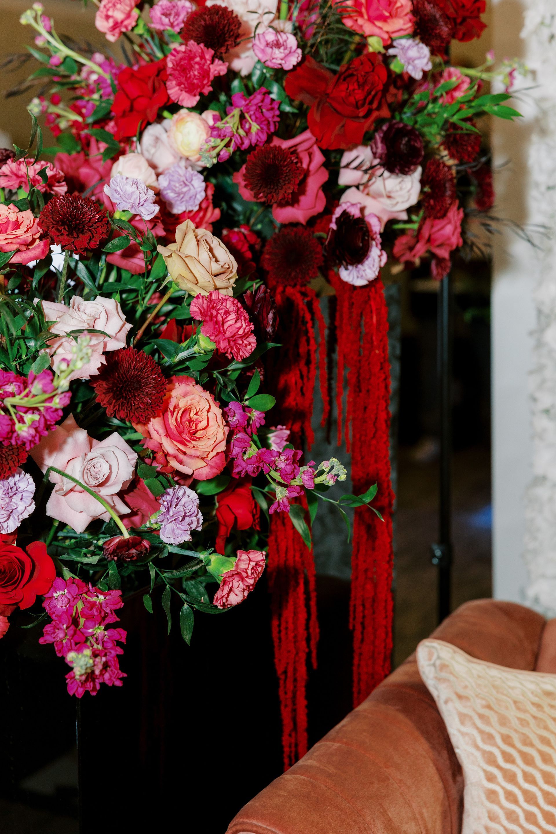 Large, vibrant flower arrangement in shades of red and pink, with red fringe accents, next to a brown couch.