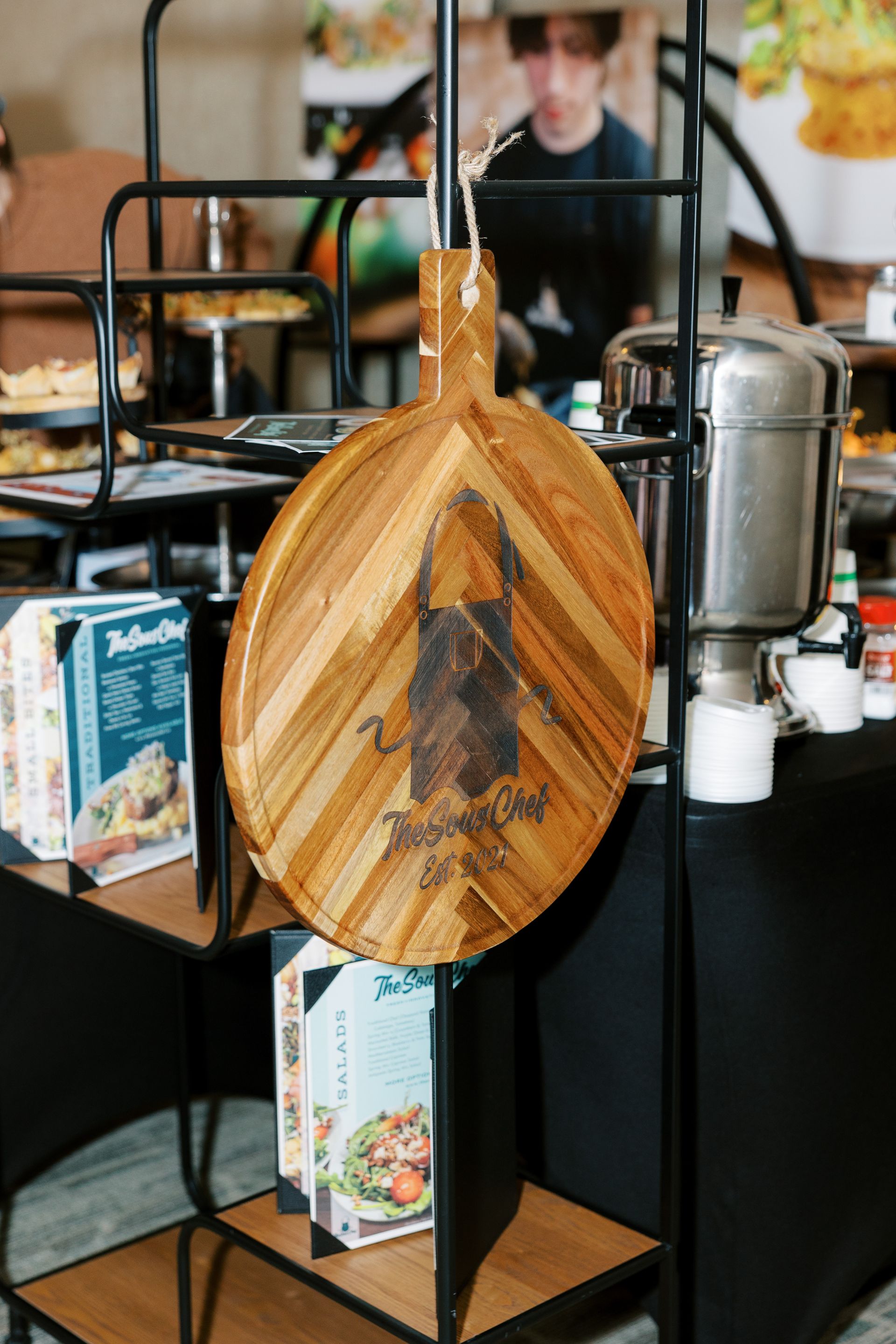 Wooden cutting board with text, part of a display. Person and food in background.