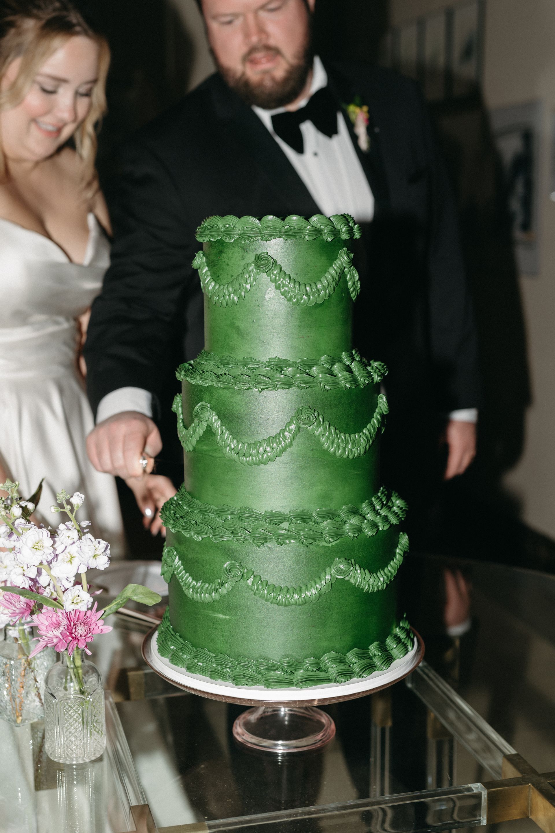 Couple cutting a three-tiered green cake at a wedding. The groom wears a tuxedo, and the bride a white dress.