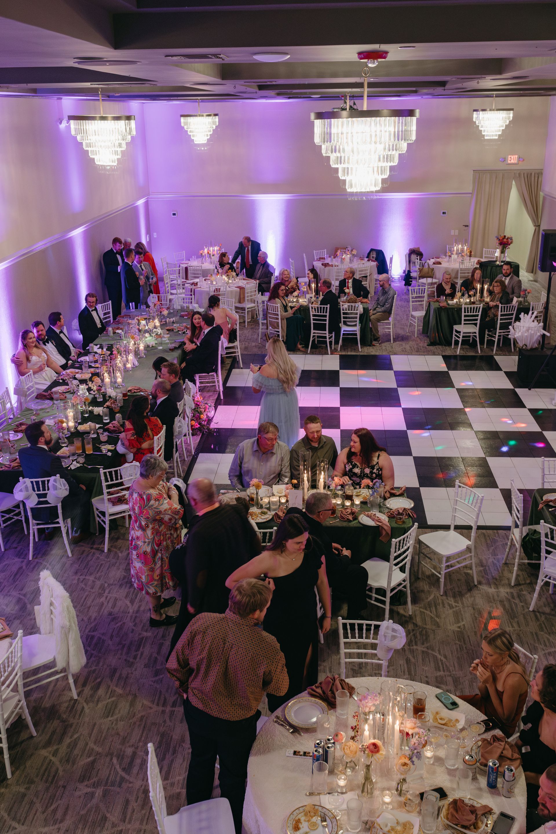 Wedding reception: guests at tables, checkered dance floor, chandeliers, purple lighting.