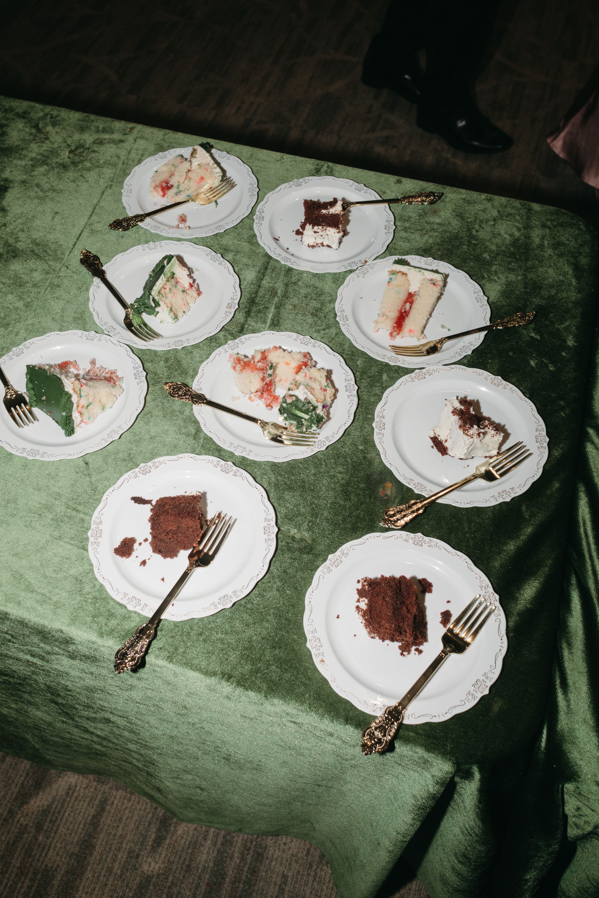 Plates of cake on a green tablecloth, with ornate forks. Black shoe and dress visible in the shot.