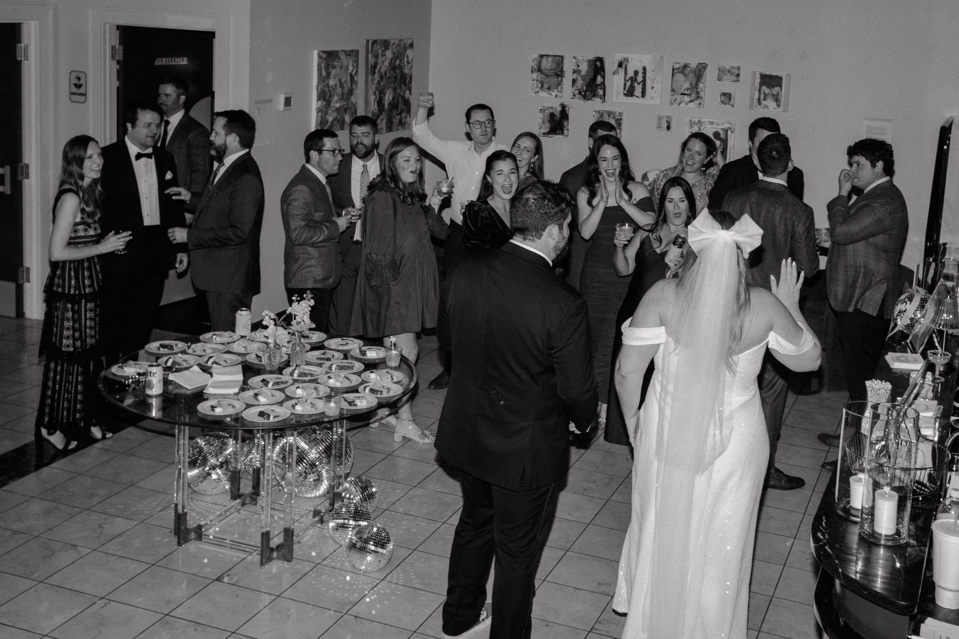Wedding reception: Guests in formal attire, bride in a veil, socializing in a room with a bar and food table.