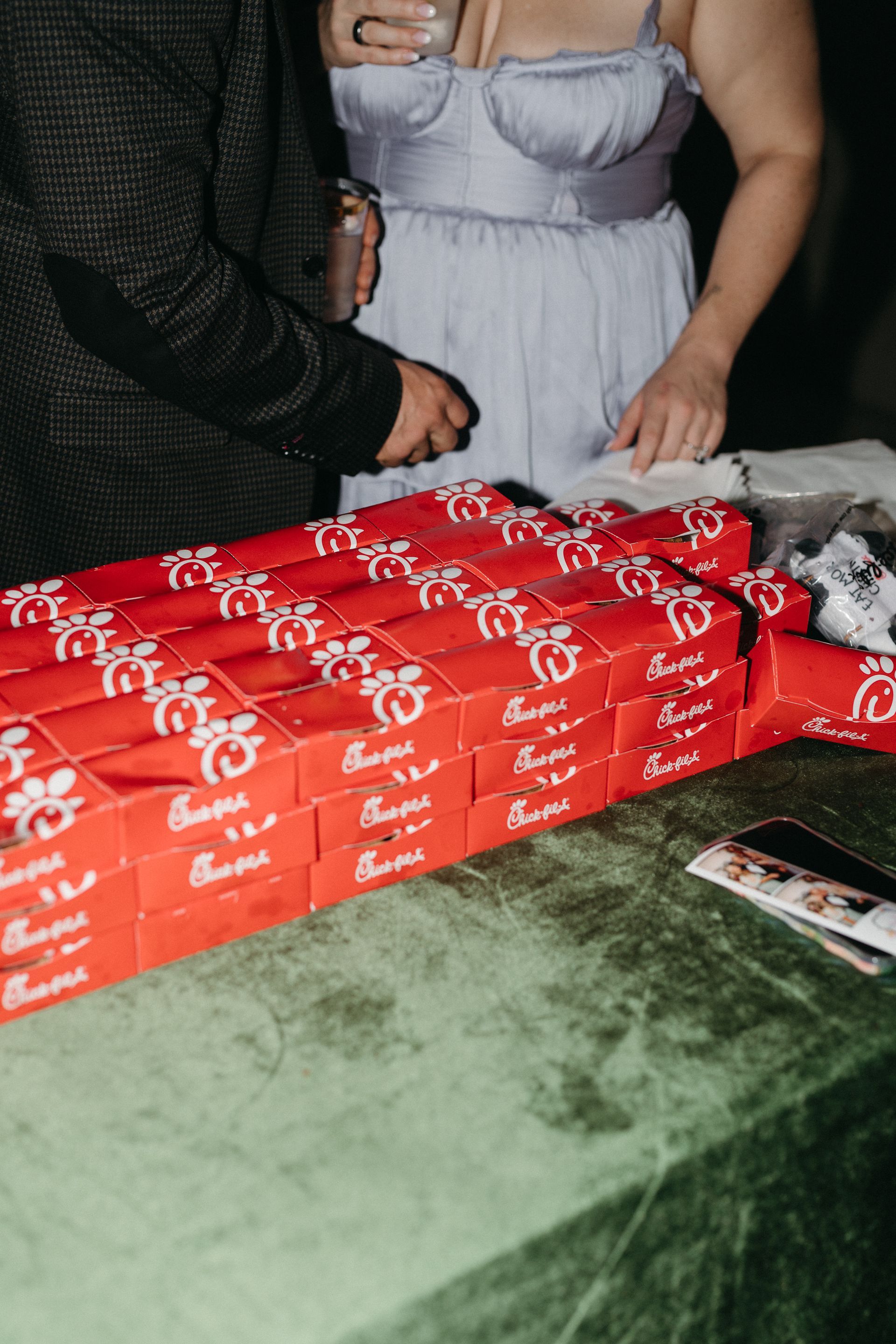 Red Chick-fil-A boxes stacked on a green table; people's hands reach for them.