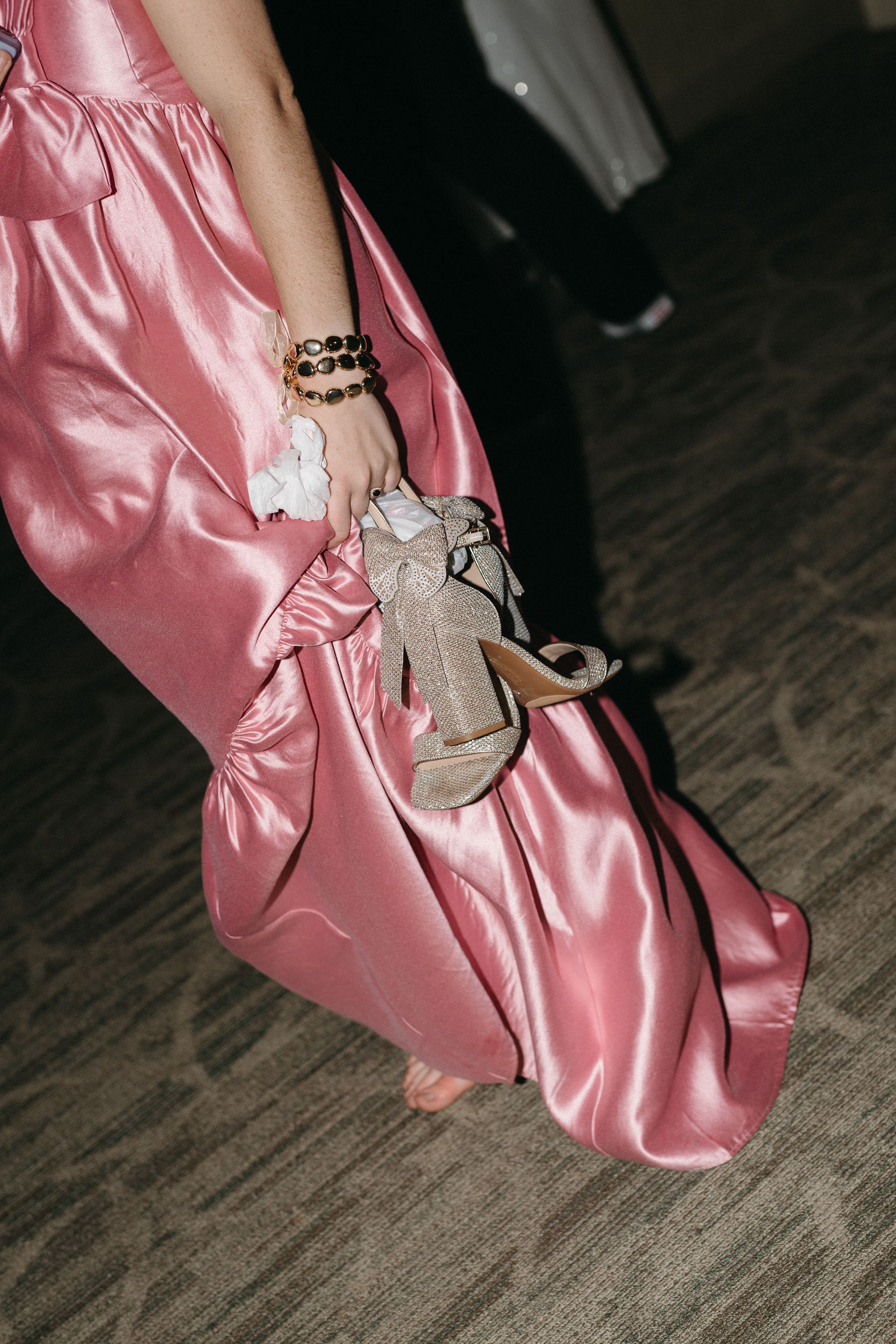 Person in a pink satin dress holding silver heels, walking in a carpeted room.