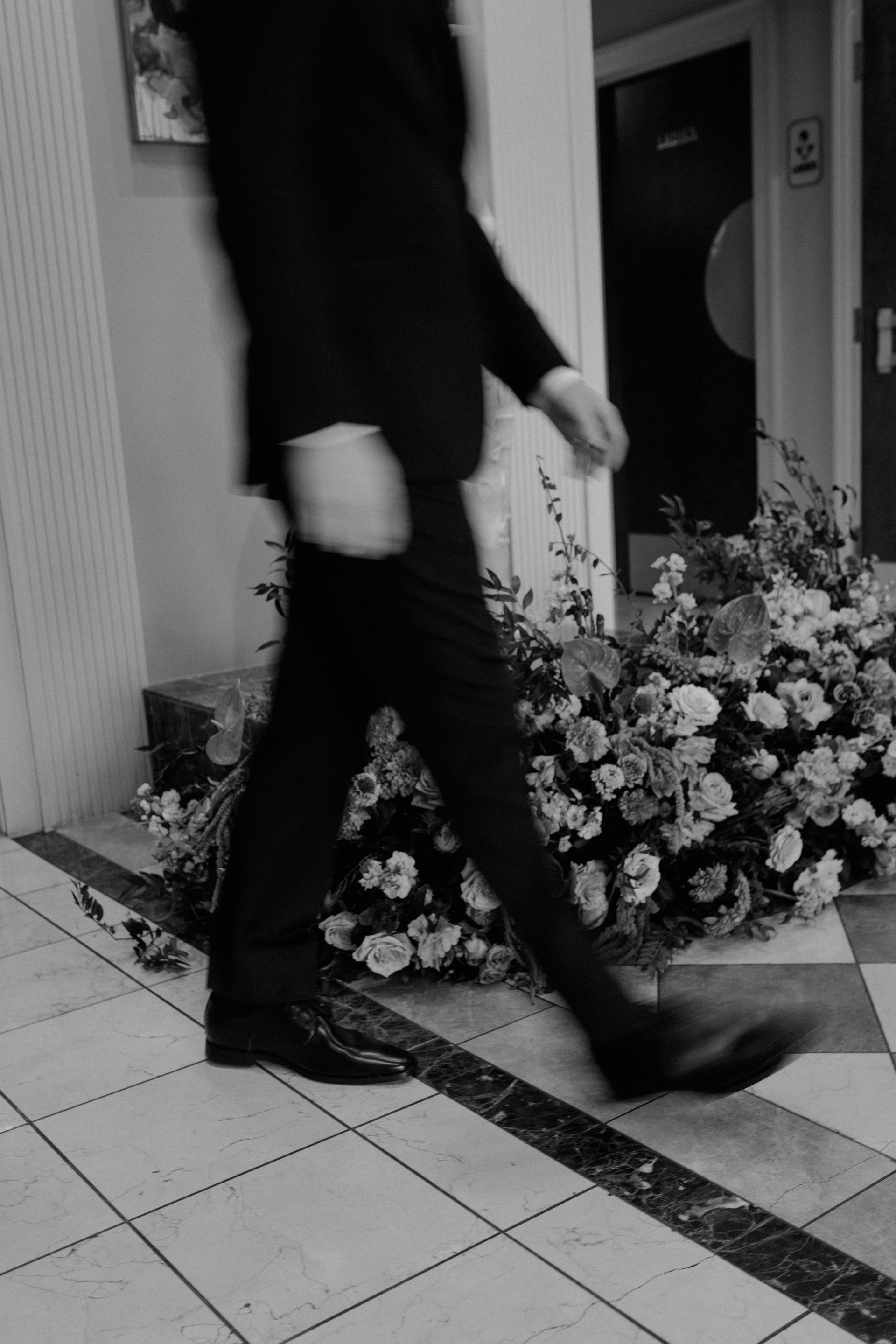Person in suit walking past floral arrangement; indoor setting.