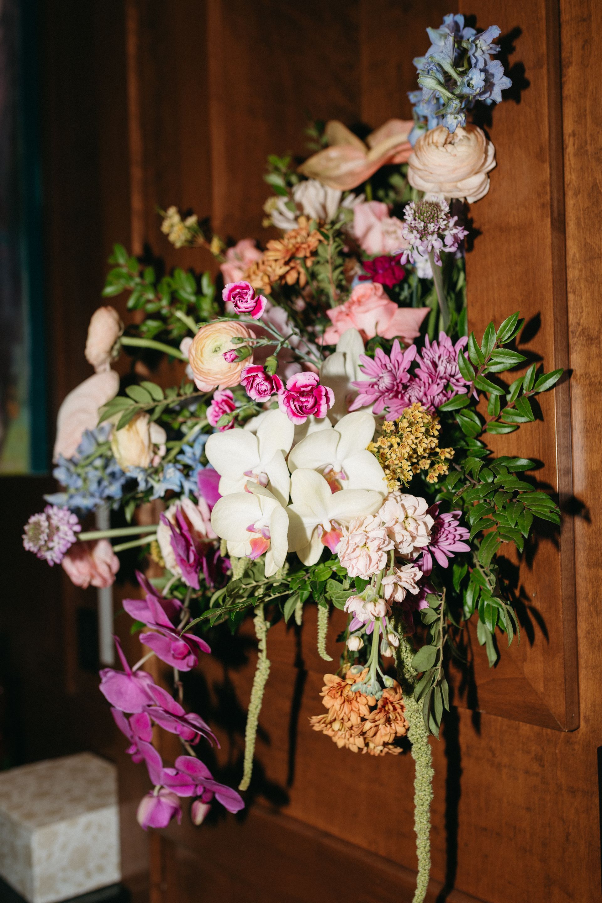 Floral arrangement with orchids, pinks, blues, and greenery on wooden paneling.