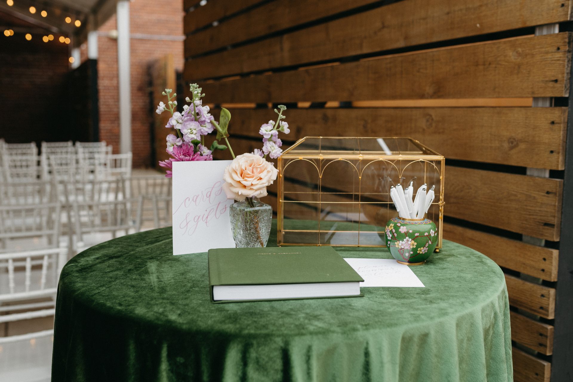 A green velvet-covered table with wedding guest book, floral arrangements, and card box. Ceremony chairs in background.