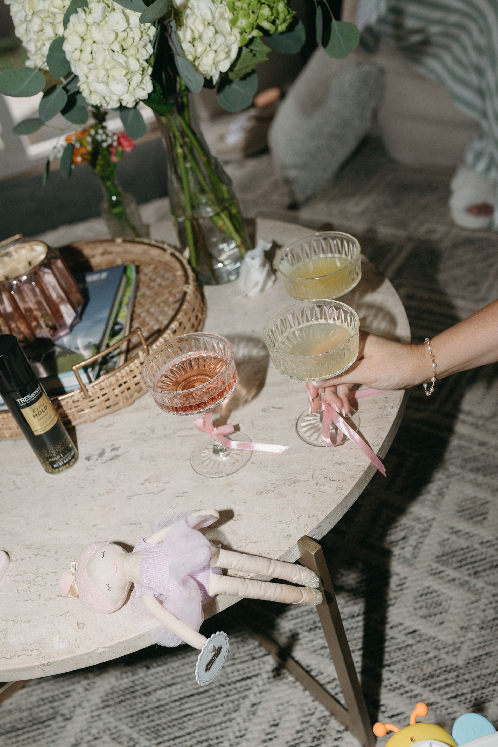 Round table with drinks, flowers, and a doll. Someone reaches for a glass.