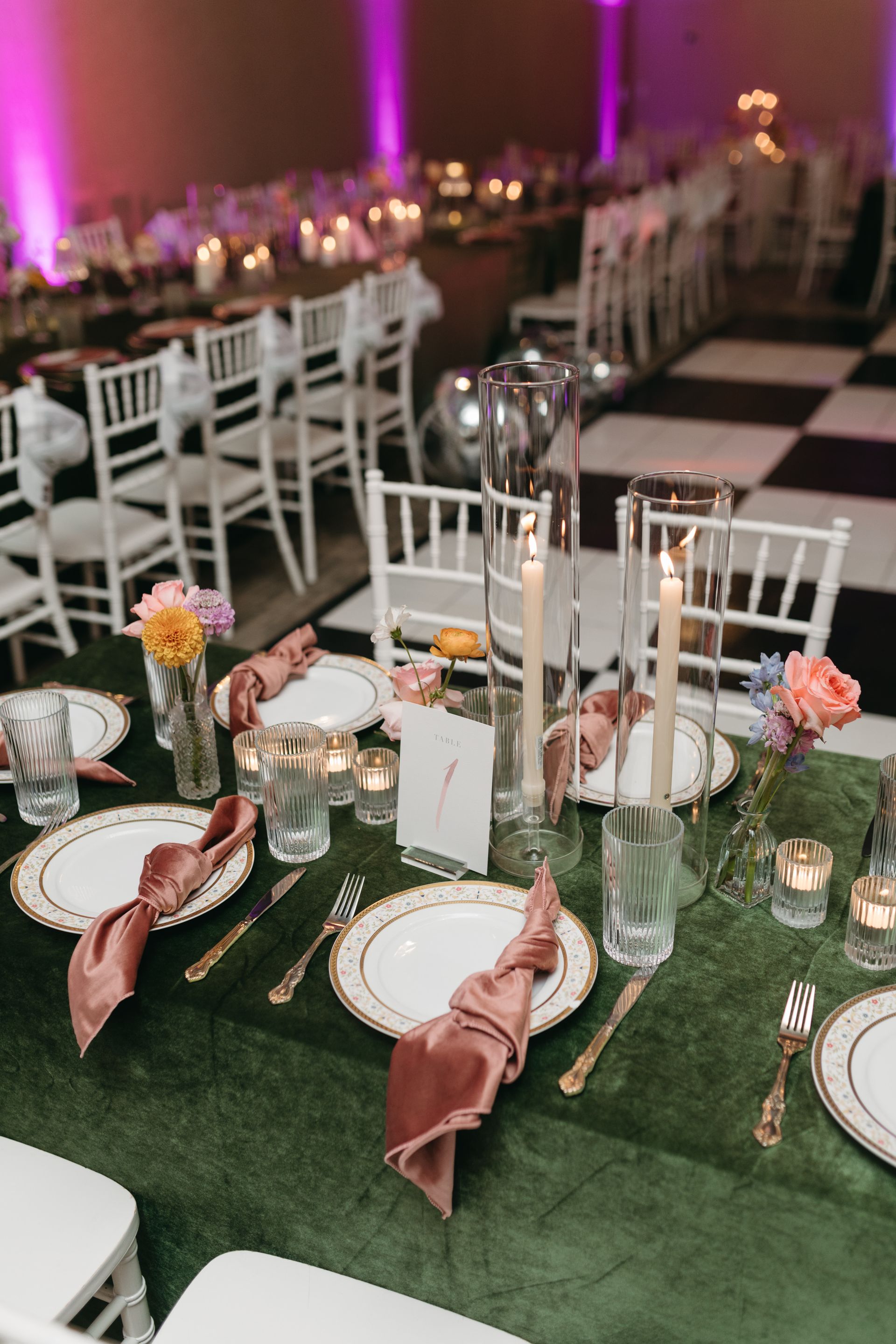 Elegant table setting with gold-rimmed plates, pink napkins, candles, and flowers on a green velvet tablecloth.