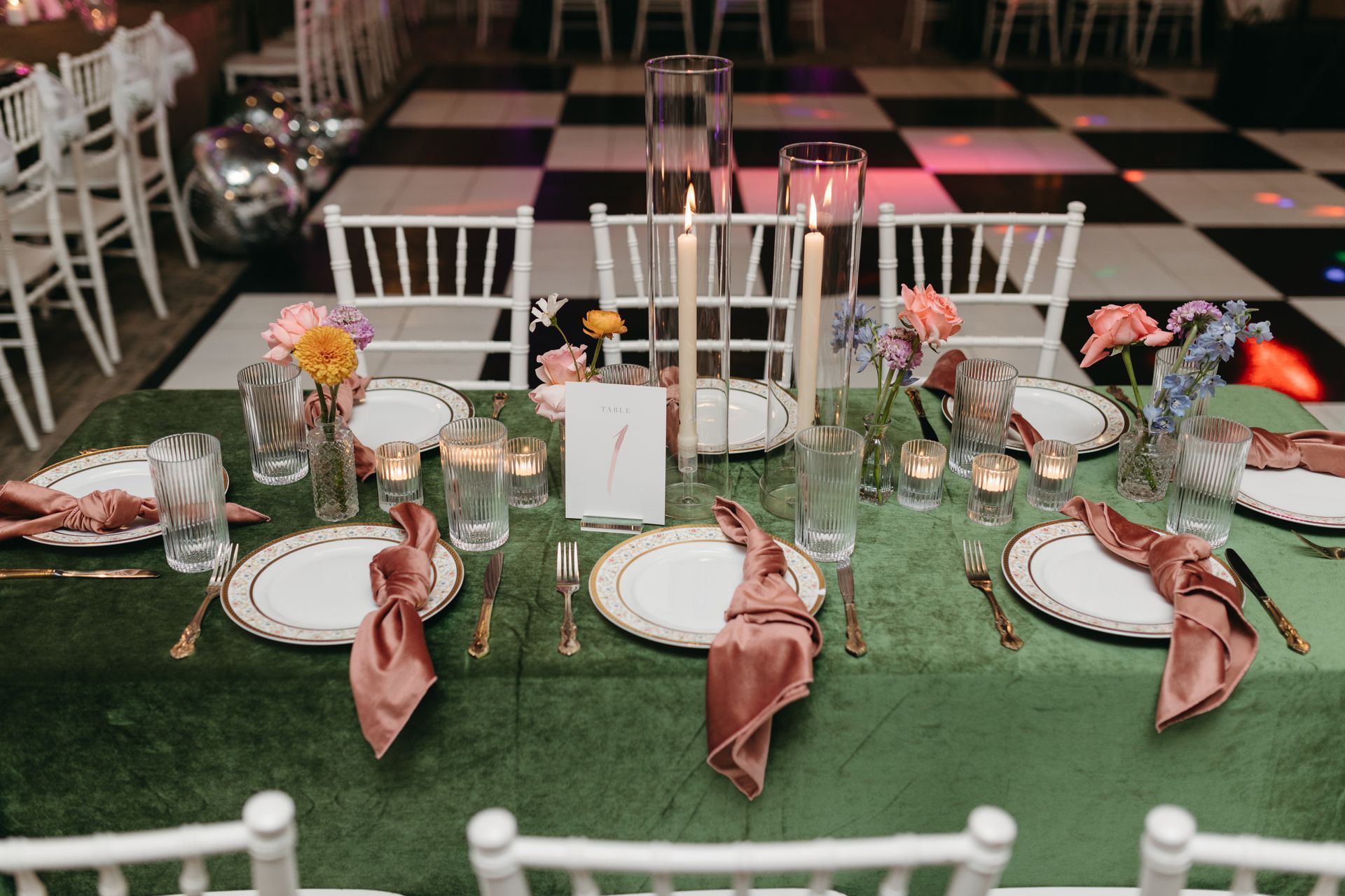 Elegant table setting with green tablecloth, gold-rimmed plates, pink napkins, candles, and flowers, white chairs on checkerboard floor.
