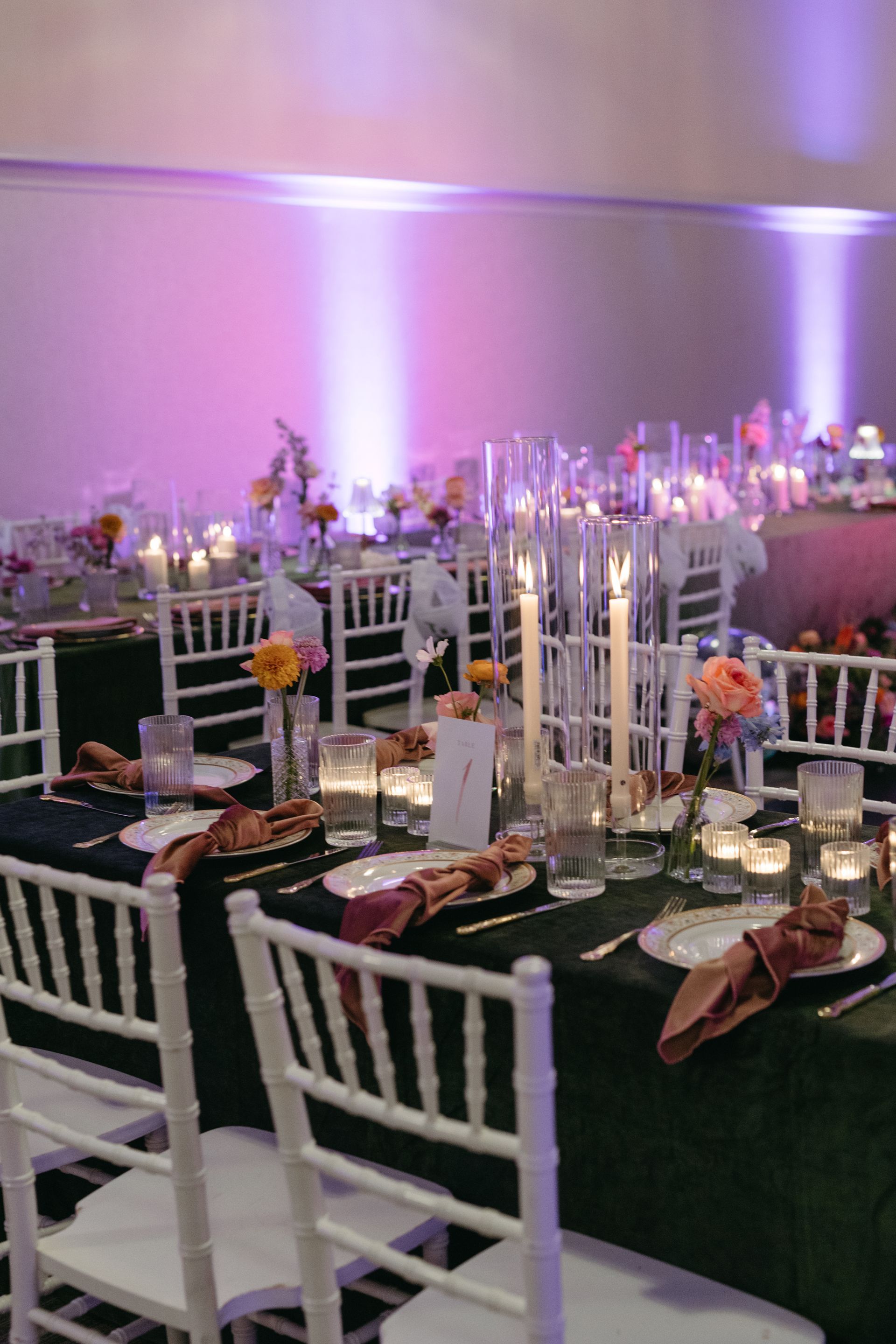 Elegant dinner table setting with white chairs, dark green tablecloth, candles, and flowers, lit with purple uplighting.