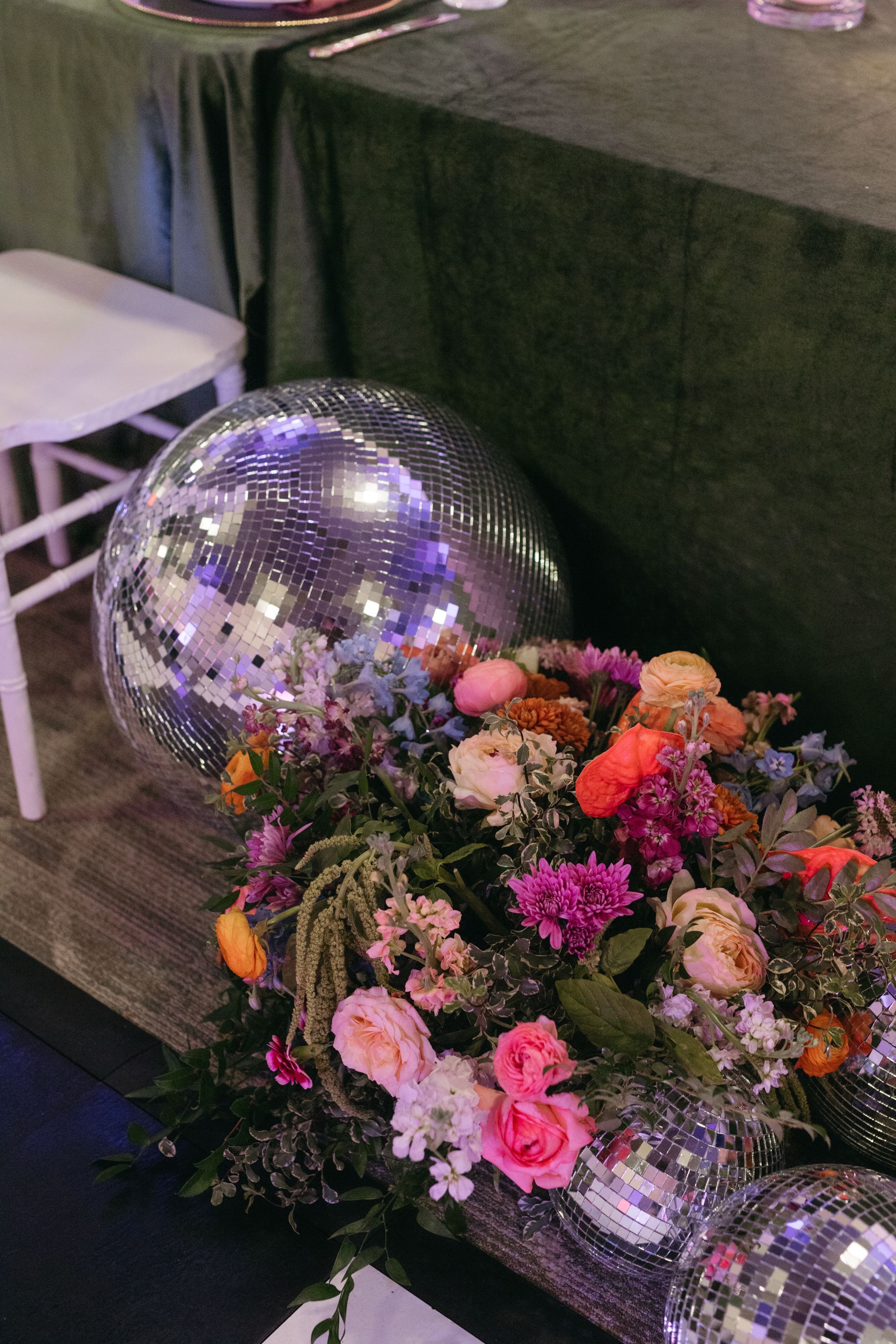 Disco balls and colorful flowers decorate a party table, with a green tablecloth and white chair in the background.