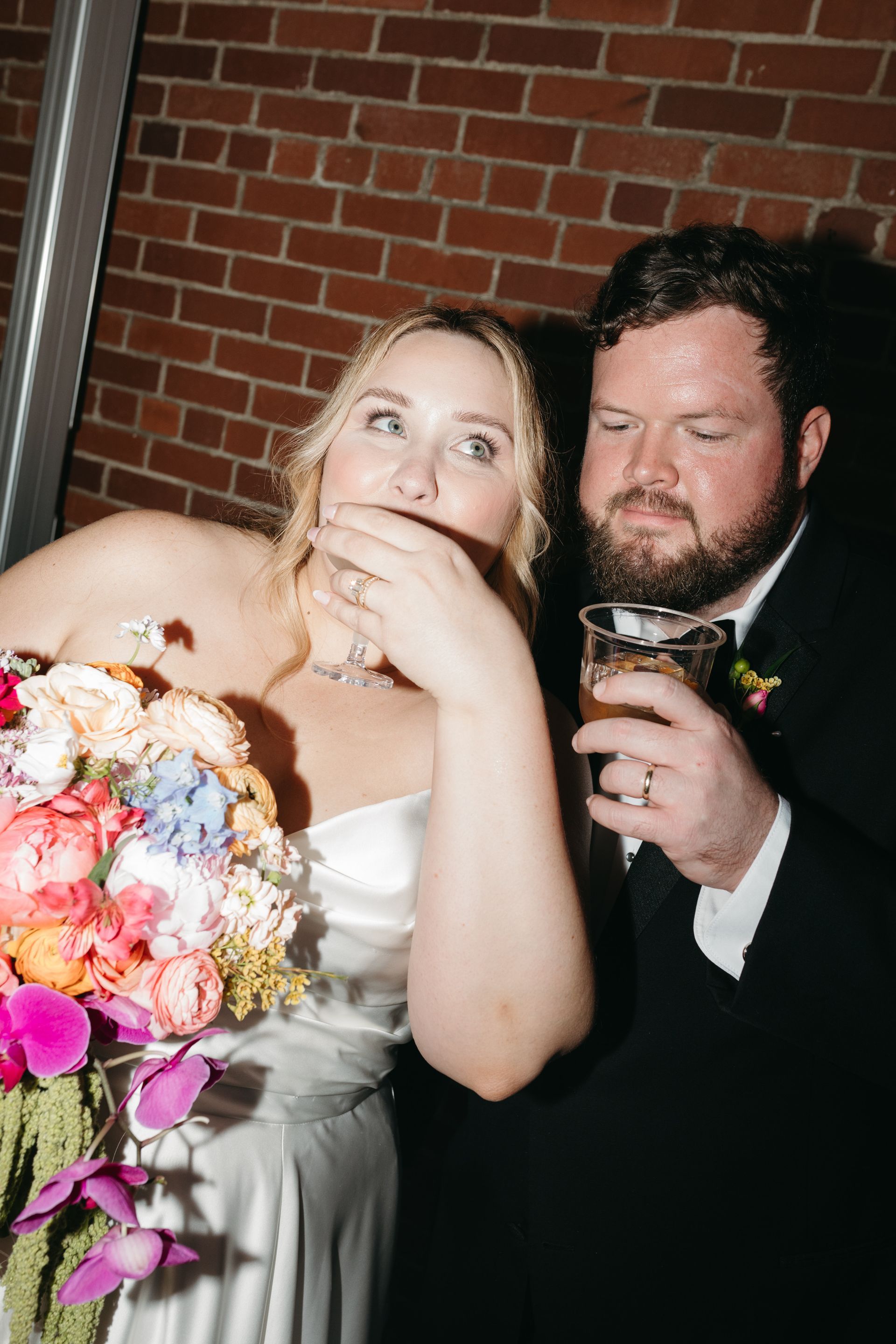 Bride eating cake, groom drinking. Wedding celebration near brick wall.