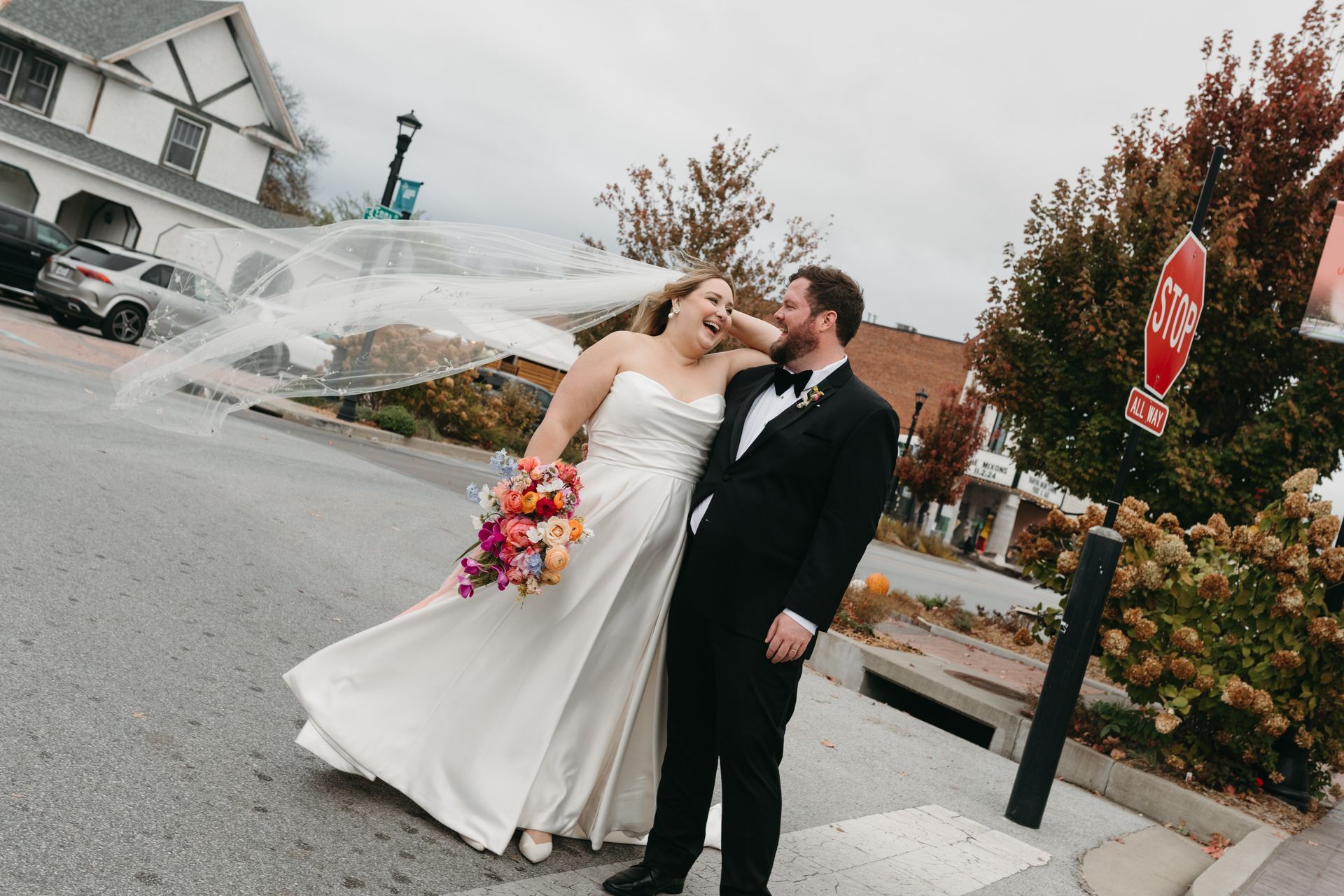 Newlyweds laughing outdoors, bride's veil blowing, holding bouquet, groom in tuxedo. Street setting, overcast day.