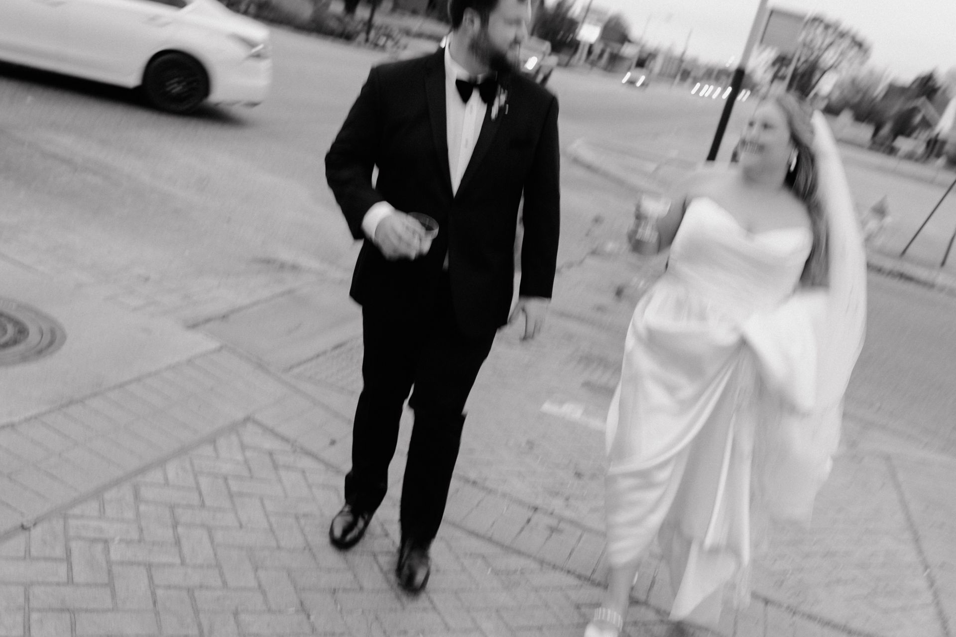Newly married couple walking on a sidewalk, groom in tuxedo, bride in white dress, holding a drink and veil flowing.