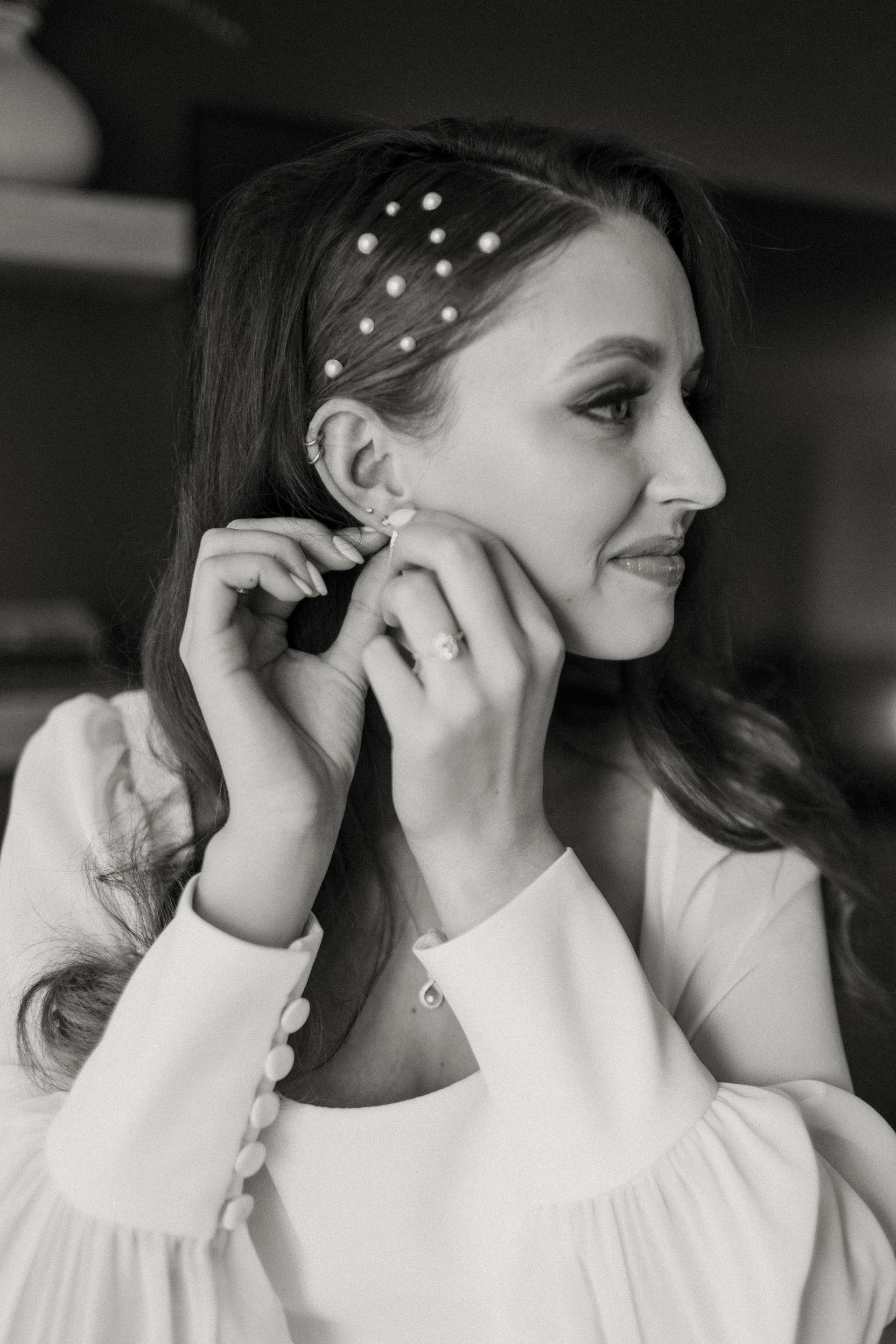 Woman putting on earring, wearing a white dress and hair adorned with pearls.