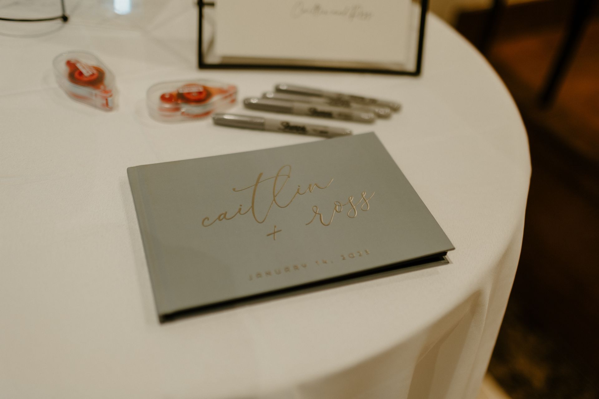 Gray guest book with gold lettering, pens, tape, on a white tablecloth.