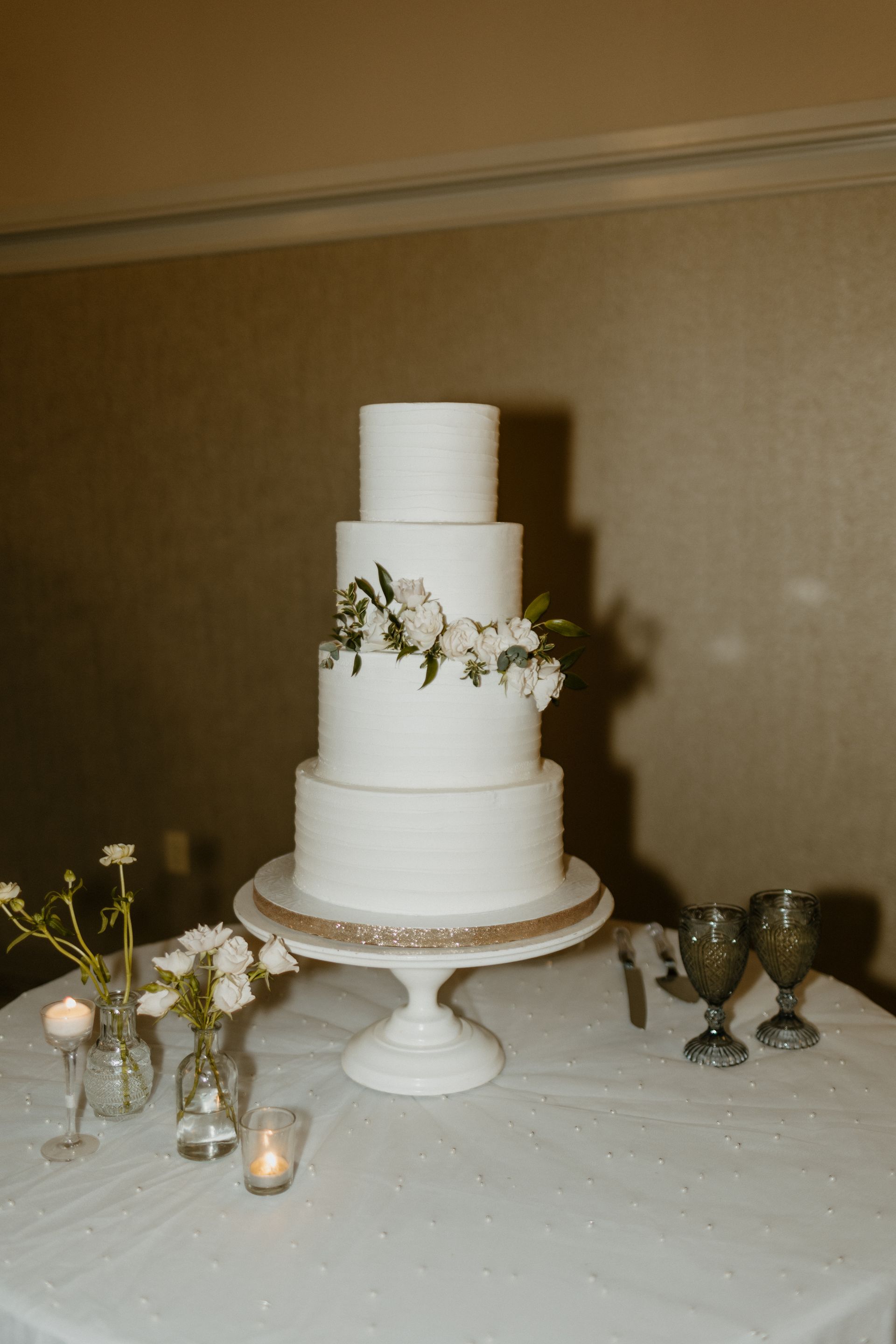 White wedding cake on a pedestal with floral decorations, two goblets, and small vases on a table.
