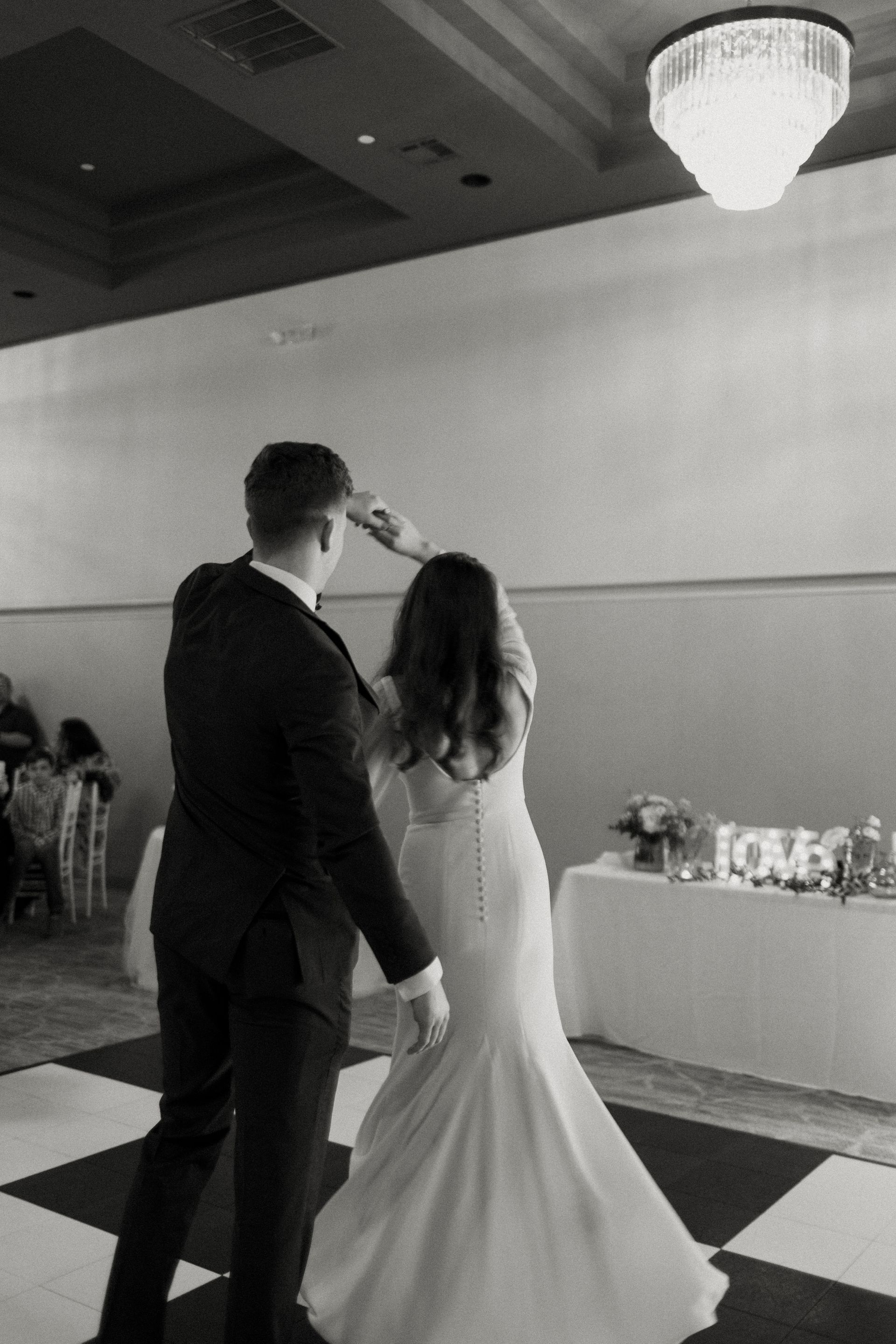 Newlyweds dance at a wedding reception on a checkered floor, with a table and chandelier in background.