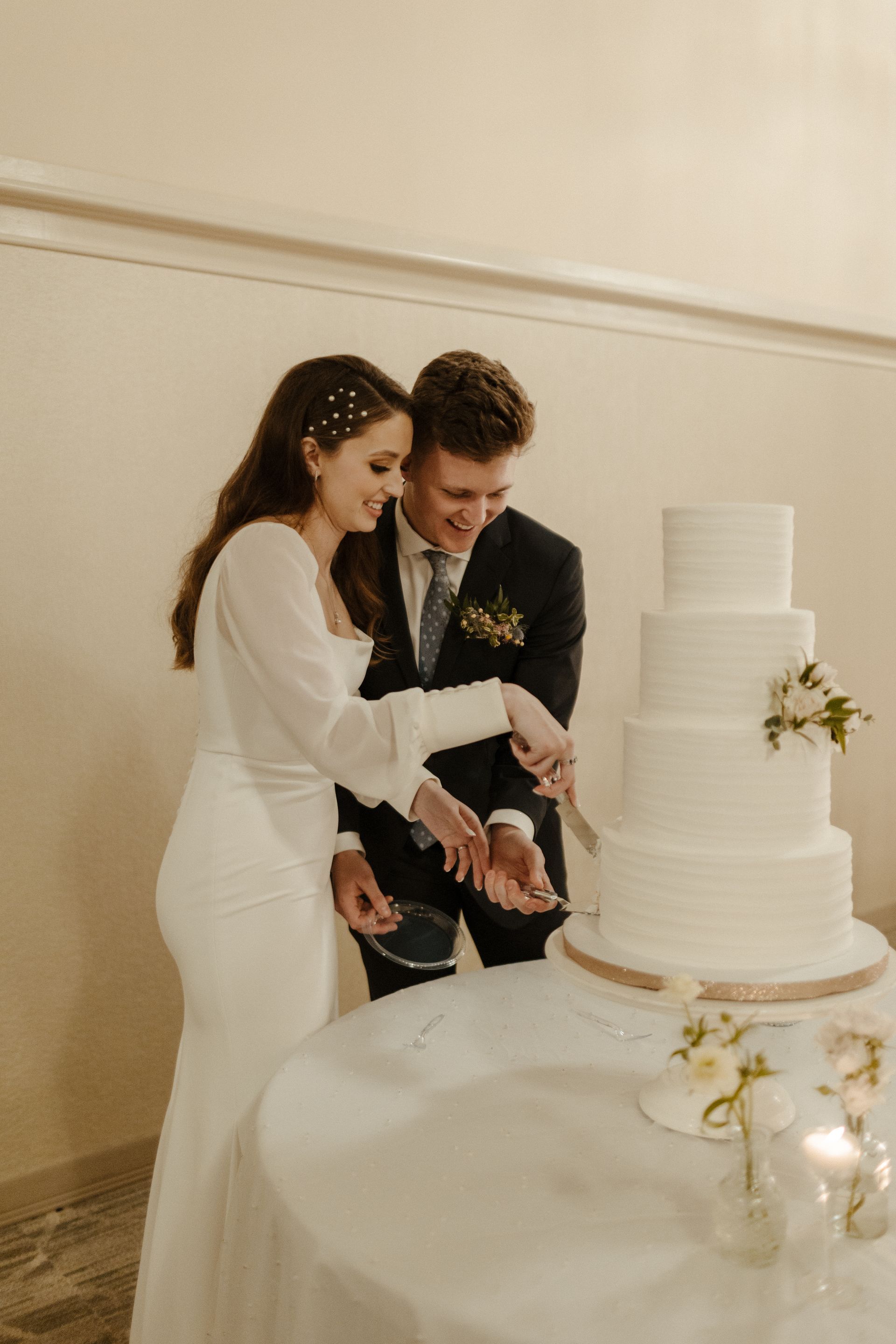 Bride and groom cut a tiered white wedding cake. They smile in a well-lit room.