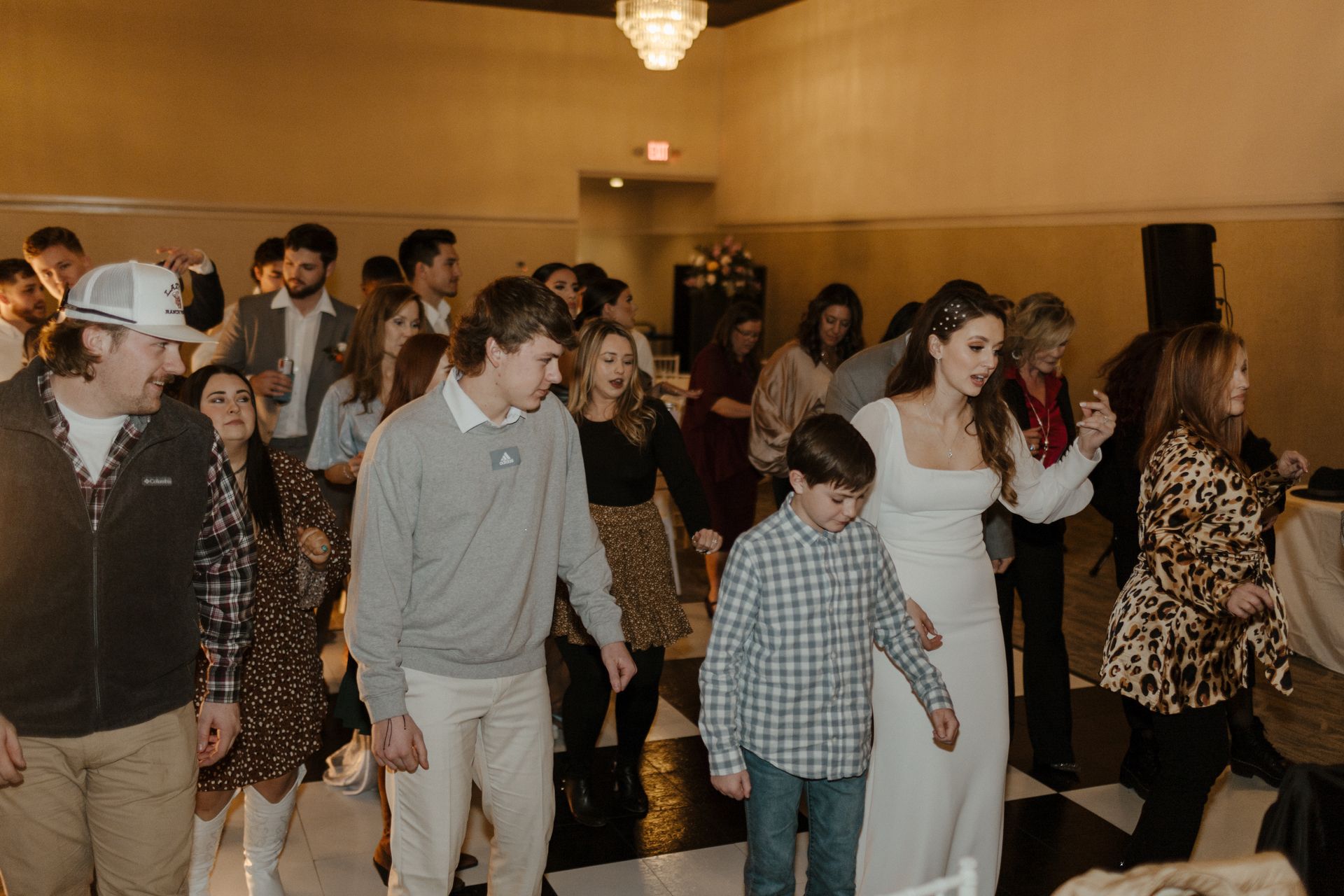 People dancing on a checkered dance floor at a wedding reception; bride leads the group.