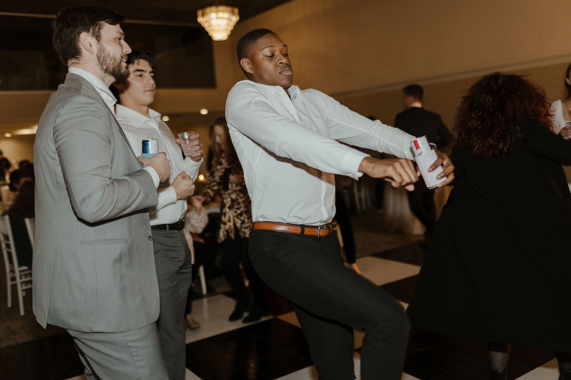 People dancing at a gathering. Man in white shirt, gesturing with arms. Black and white checkered floor.