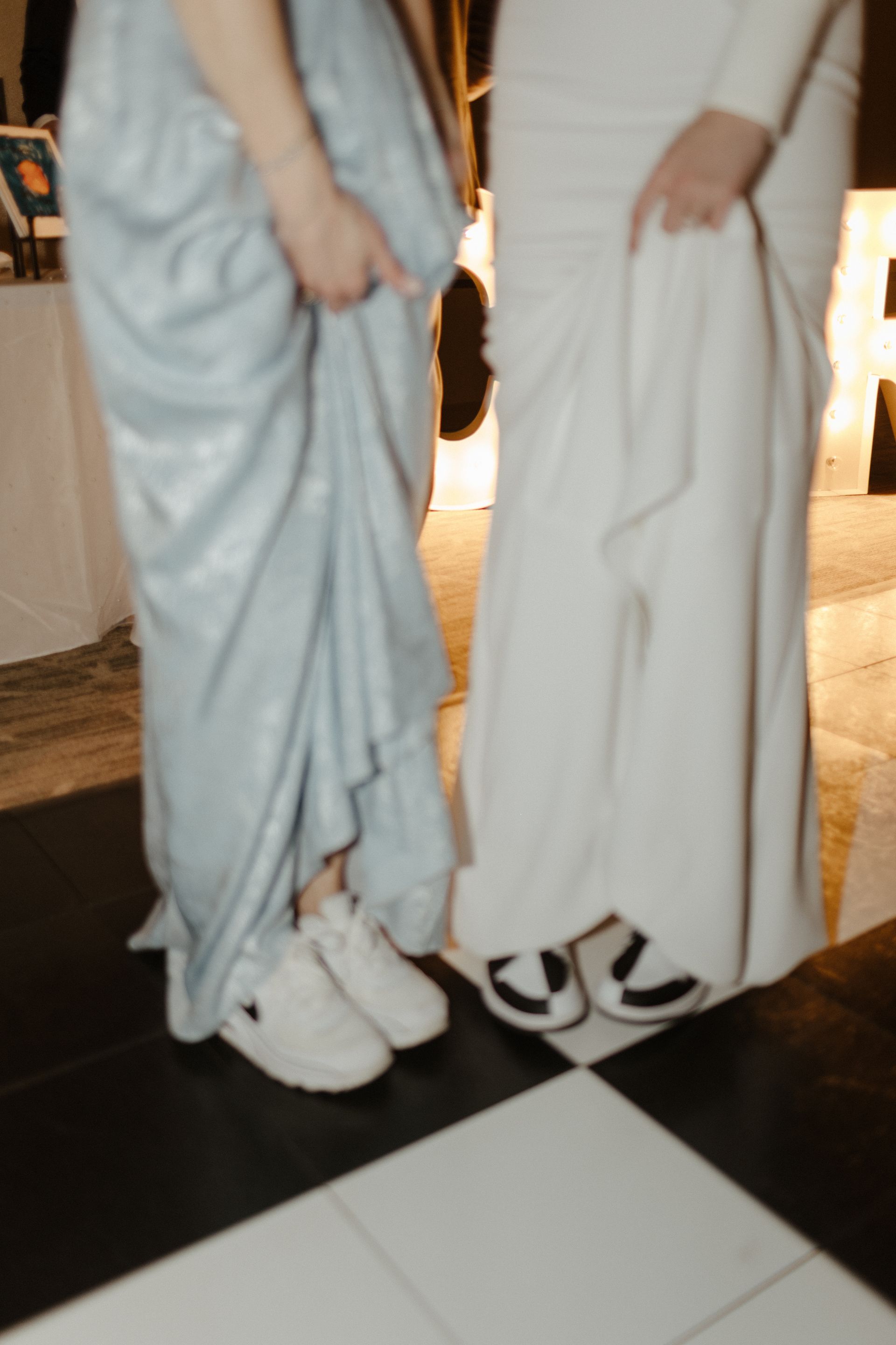 Two people in long dresses hold up skirts, revealing sneakers and sandals on a black-and-white checkered floor.
