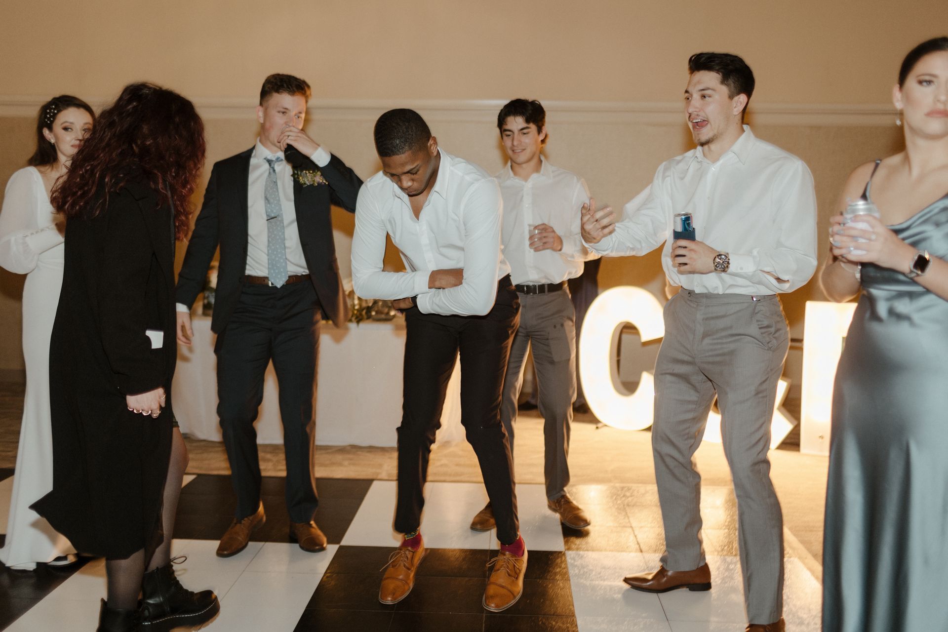 Group dancing on a checkered dance floor in a well-lit room. Some are smiling, others are holding drinks. Large illuminated letters are in the background.