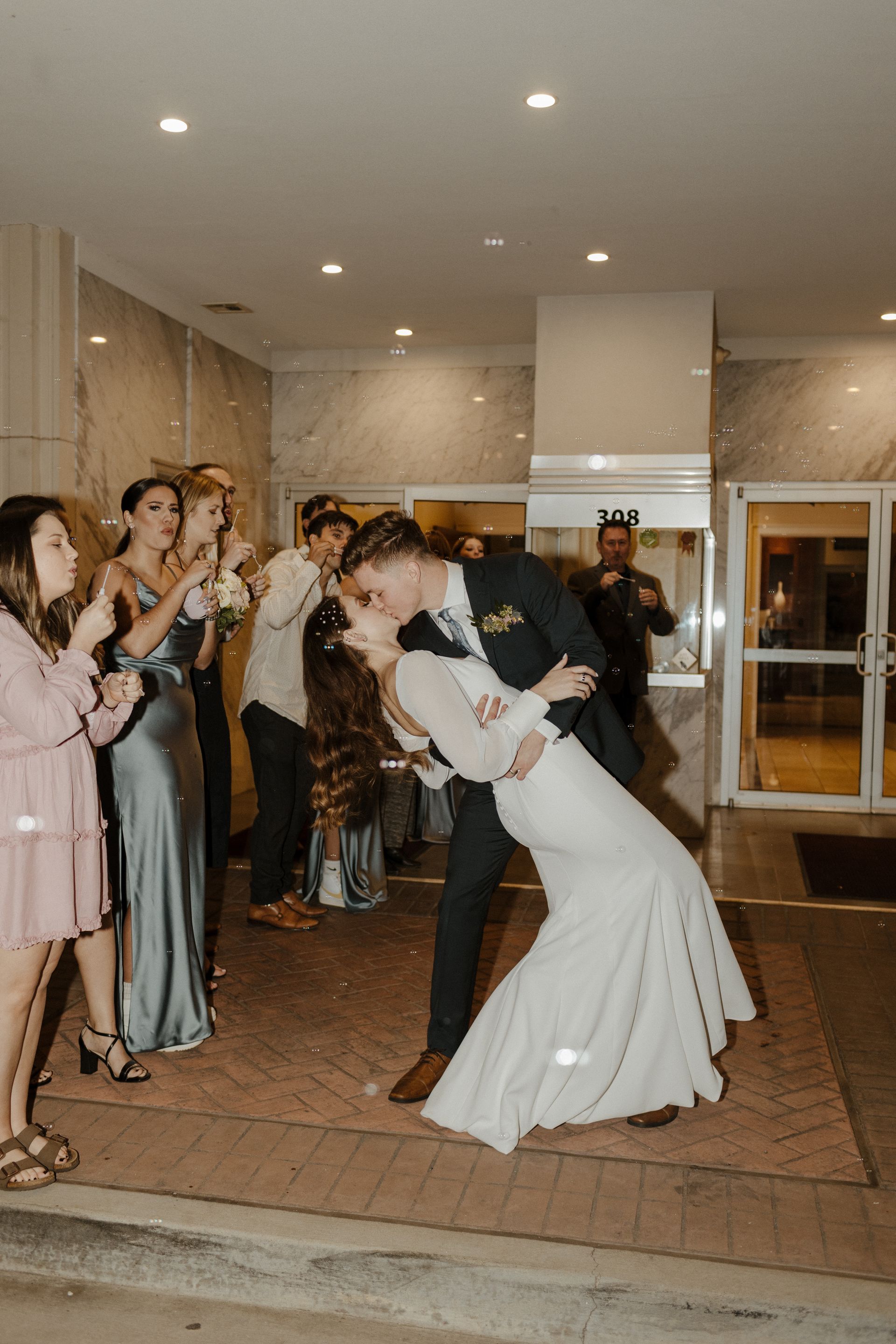 Women in a wood-paneled room, conversing and holding drinks near a floral arrangement, with a window and bar area in view.