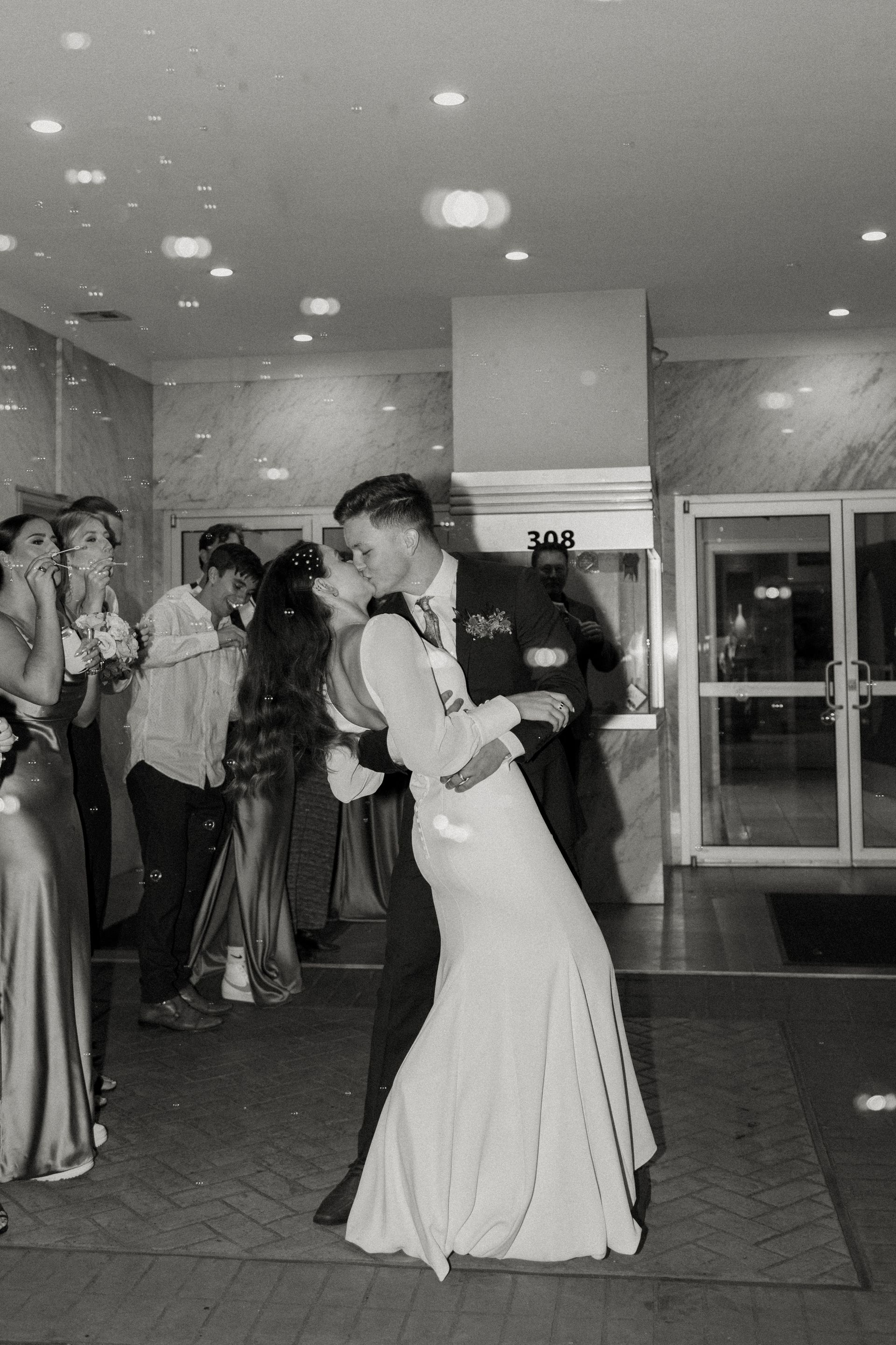 Couple kissing while dancing at a formal event, with onlookers in the background. Black and white.