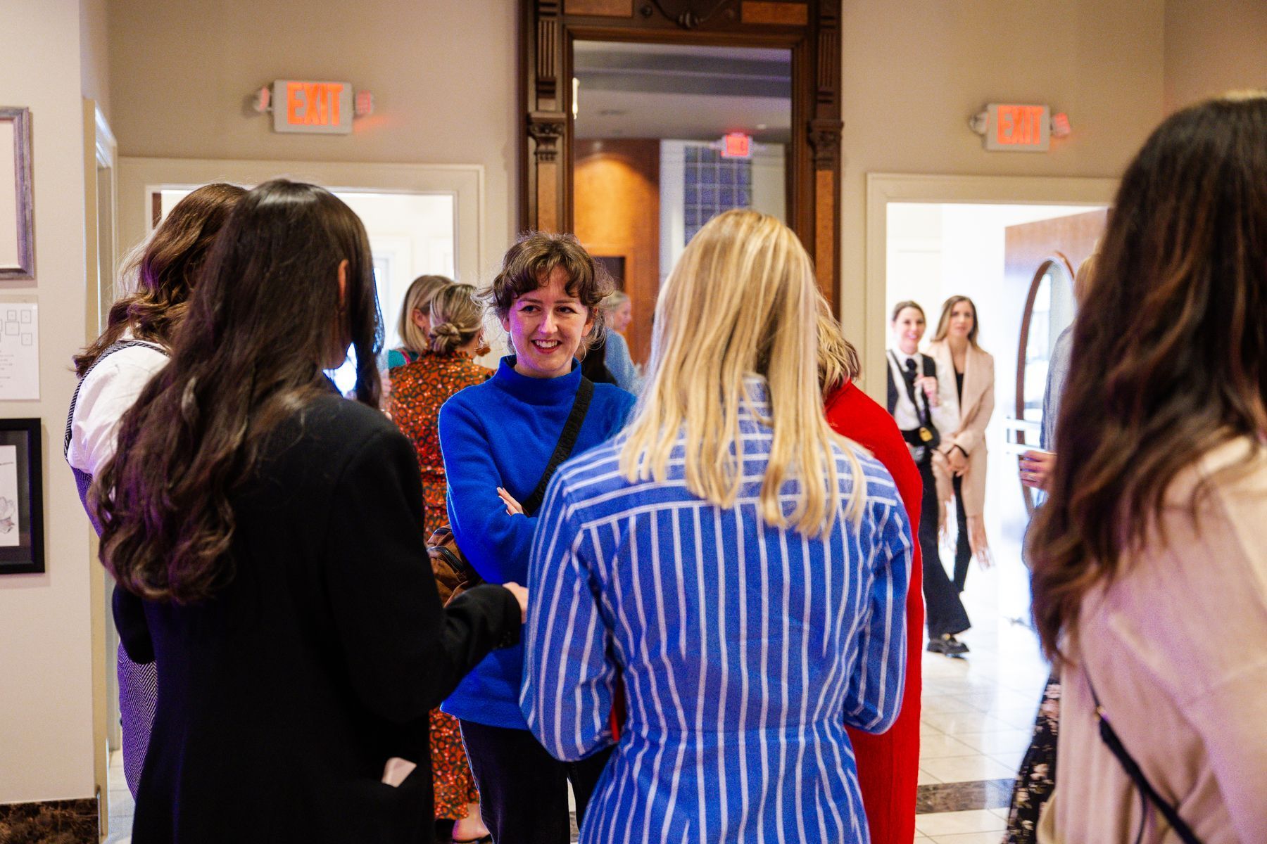 People in hallway, some talking. Woman in blue smiling, blonde in striped shirt, other people in background.