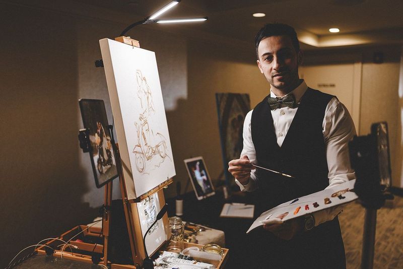 Artist painting at an easel, holding a palette and brush, wearing a vest and bow tie, indoors.