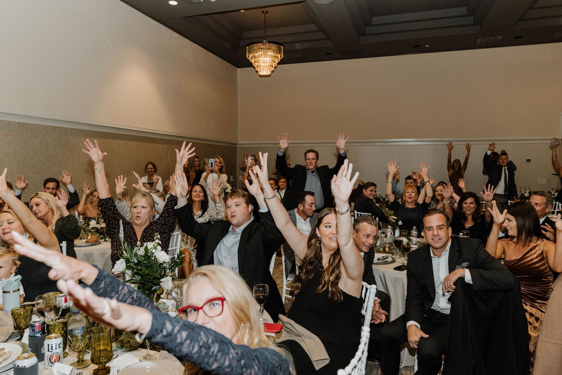 Guests at a celebration, arms raised in the air. Banquet tables with decor are in a large room.