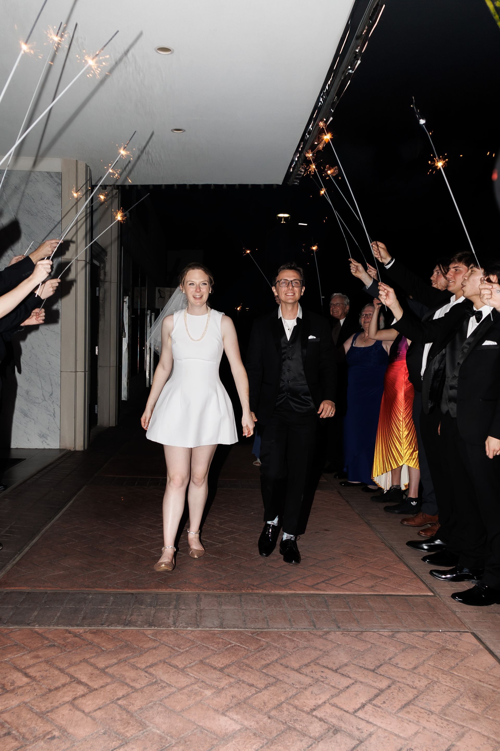 Couple exits building under sparkler archway. Night scene with formal attire and smiling faces.