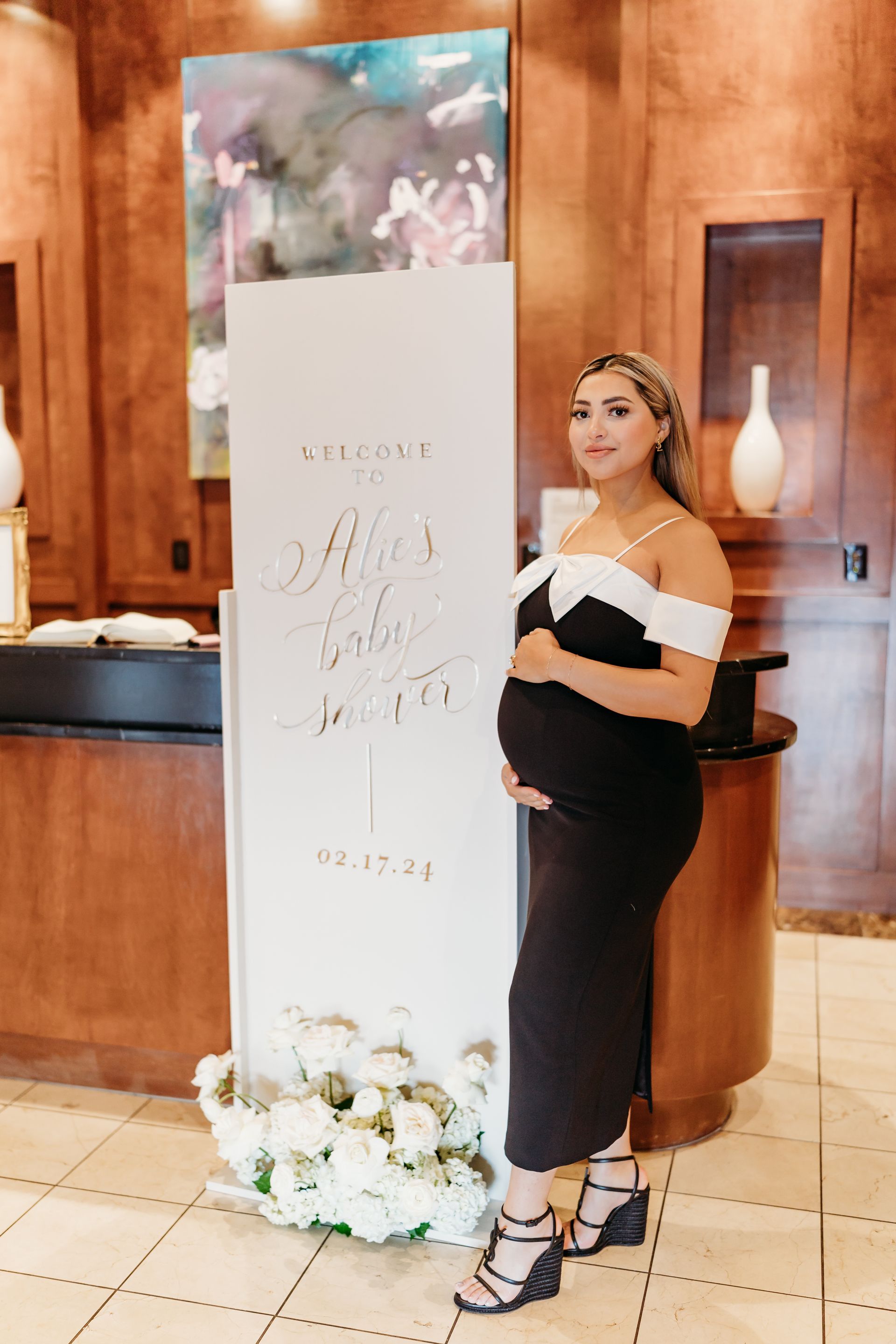 Pregnant woman in black dress poses next to a white welcome sign with floral arrangement.