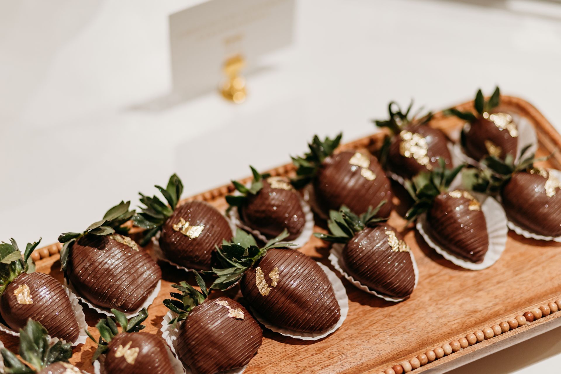 Chocolate-covered strawberries on a wooden tray with gold flakes, near a white card.