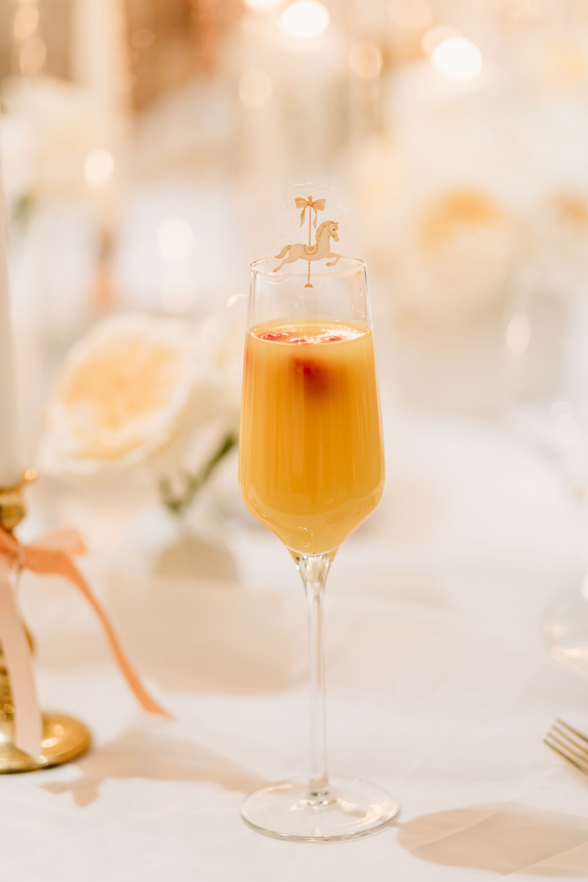 A champagne flute with an orange drink and a golden carousel horse decoration, on a white tablecloth.