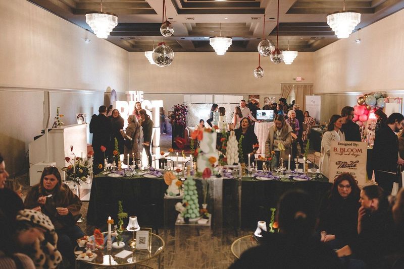 Event with tables displaying items, attendees in a large room with chandeliers and disco balls.