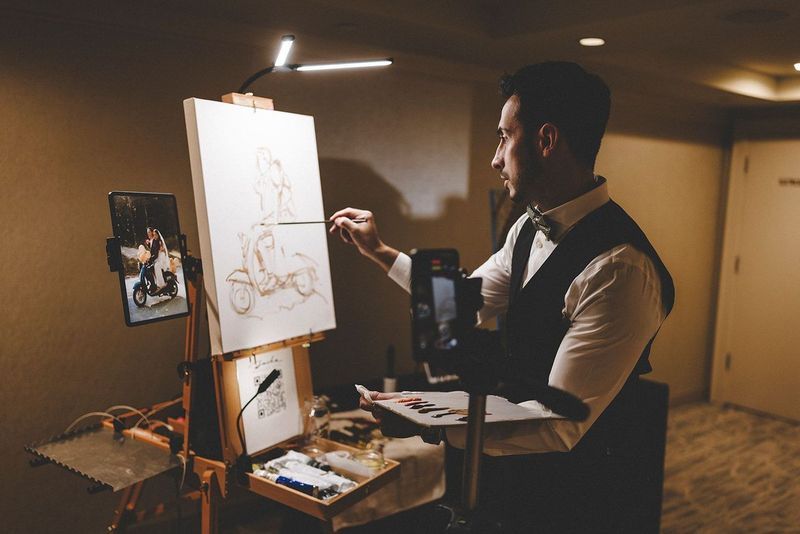 Man in vest painting on easel, lit by a lamp. Two phones record the art process in a room.