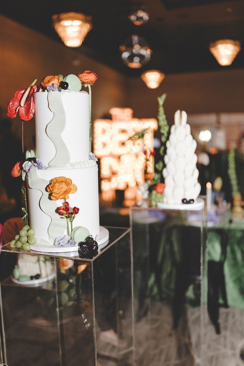 Two-tiered white cake with floral decorations on clear acrylic stand. Restaurant setting with blurred background.