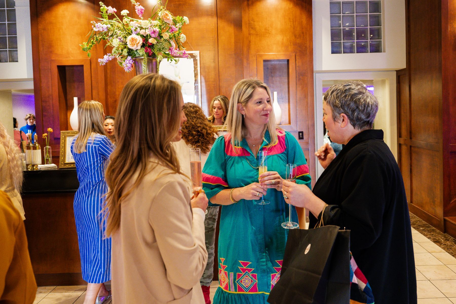 Women in a wood-paneled room, conversing and holding drinks near a floral arrangement, with a window and bar area in view.